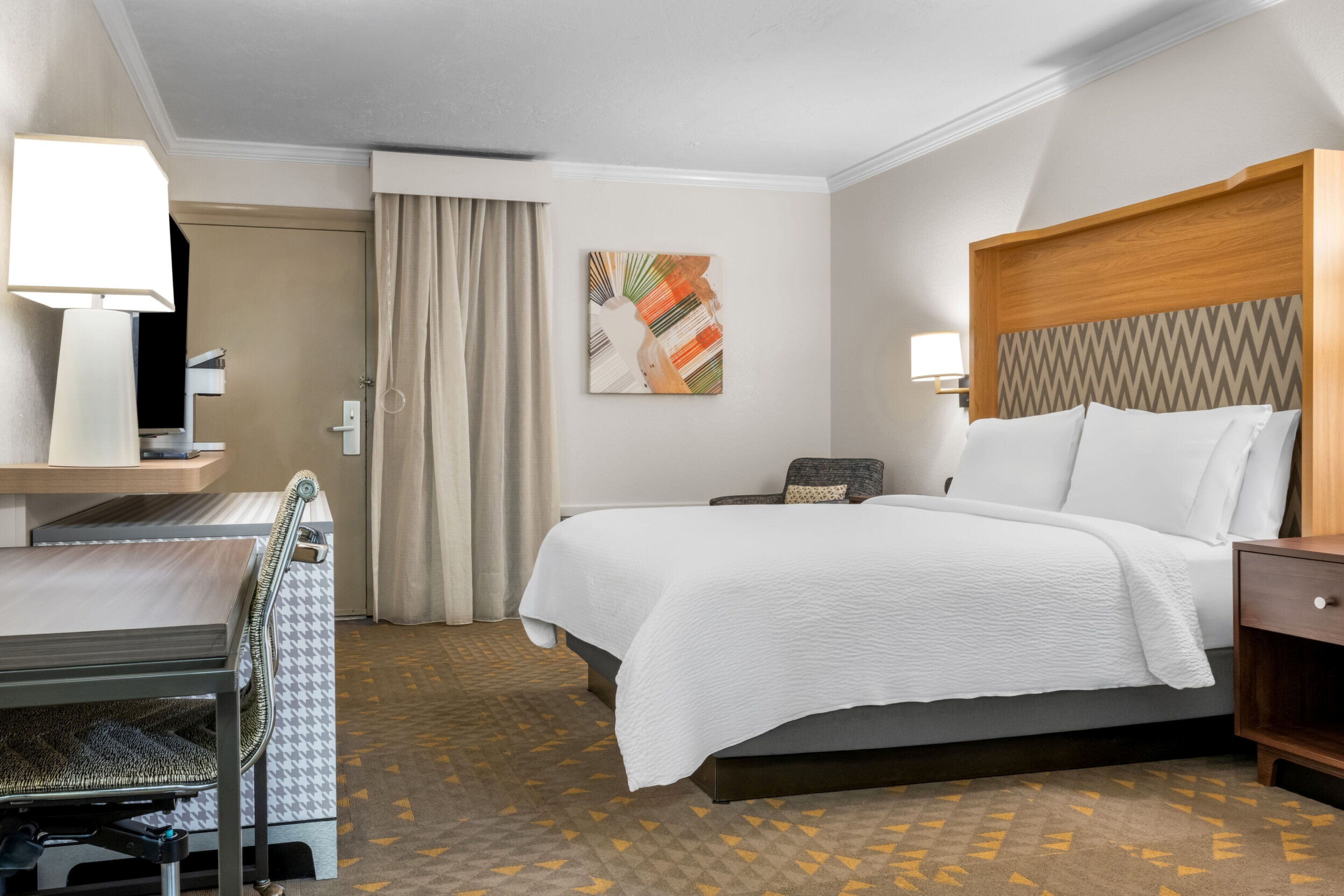 A hotel room with a bed, desk, chair, and a painting on the wall.