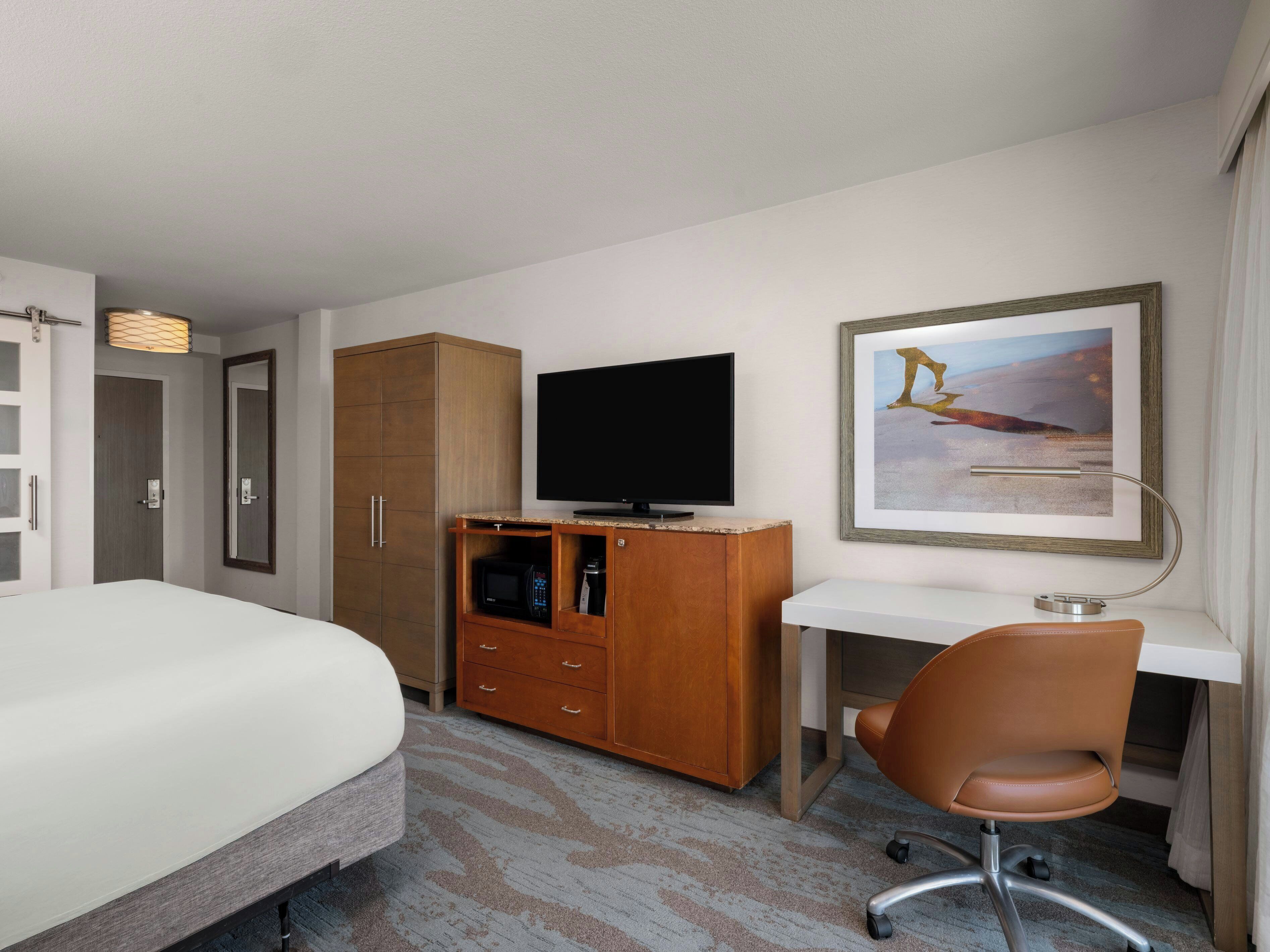 A hotel room with a bed, a TV, a desk, a chair, a wardrobe, and a framed picture on the wall.