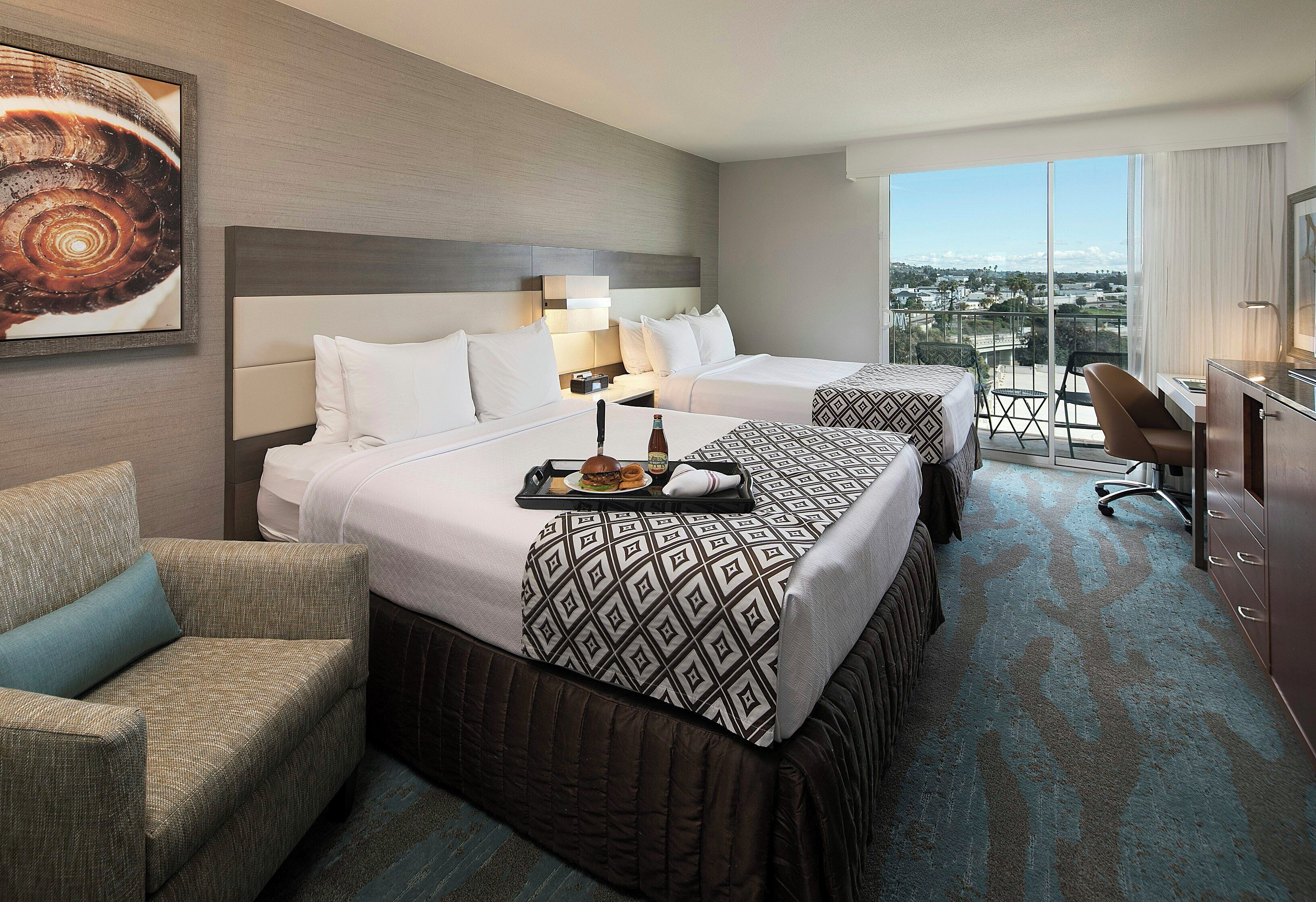 A hotel room with two beds, a sitting area, a desk, and a view of the city.