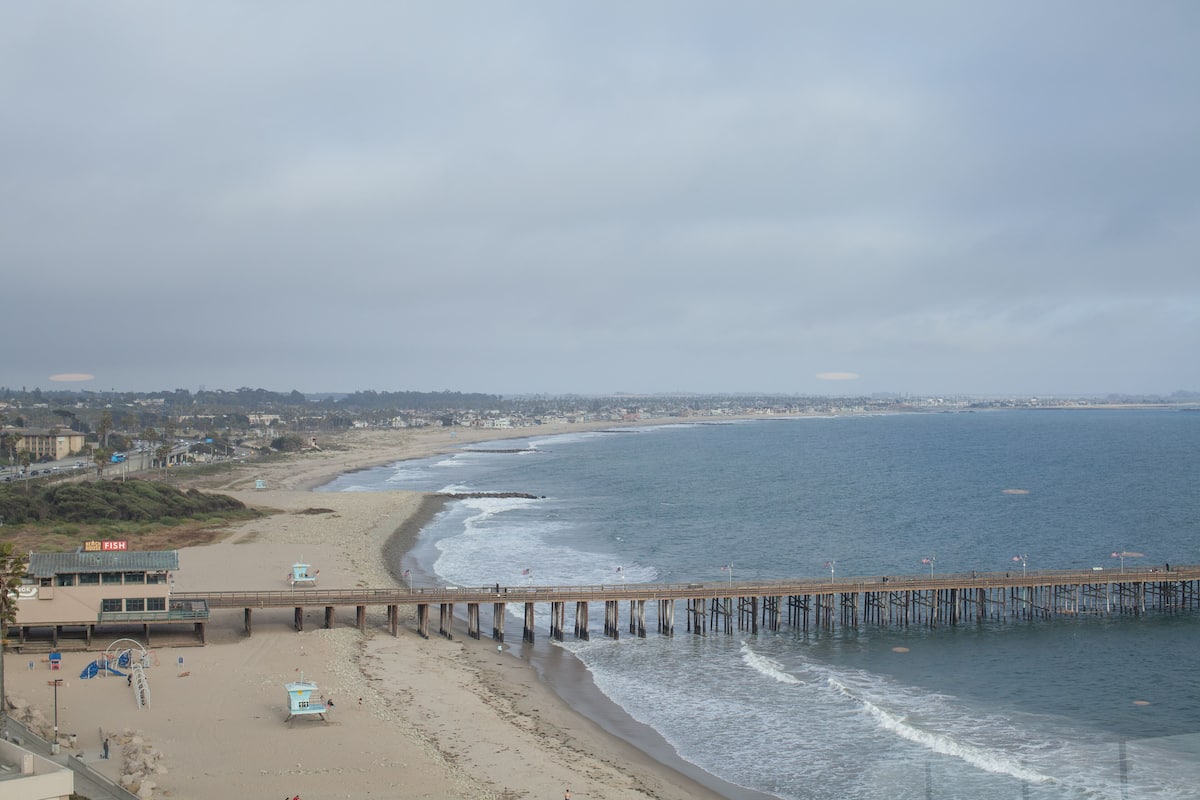 A beach with a pier extending into the water, a lifeguard tower, and a beach chair area.