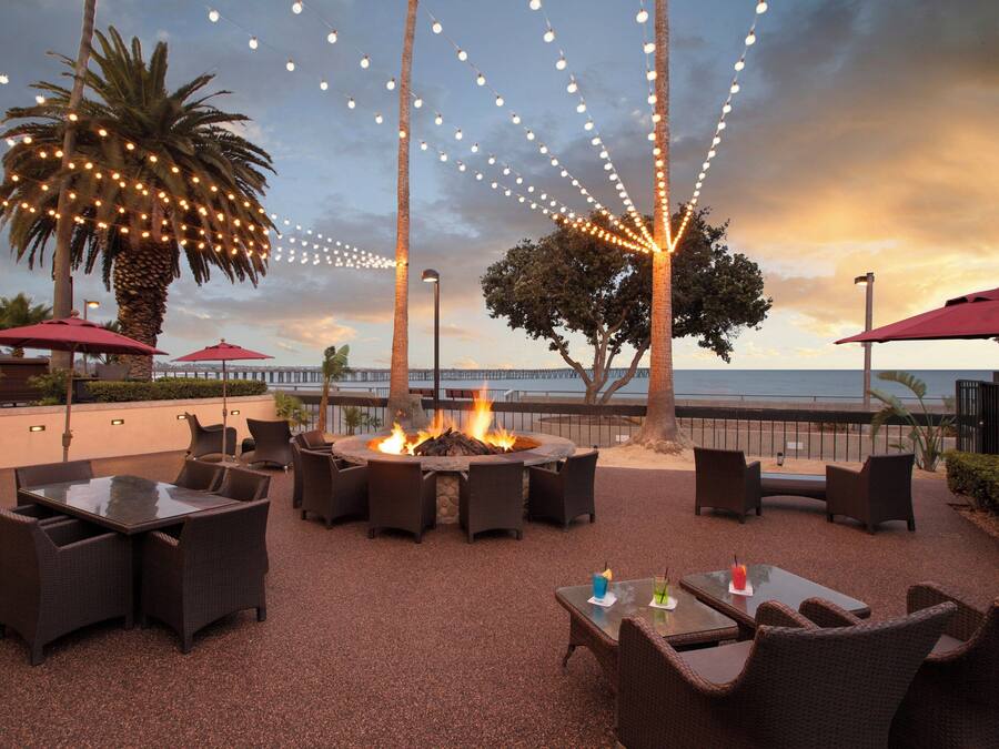 An outdoor seating area with wicker furniture, a fire pit, and string lights against a sunset backdrop.