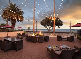 An outdoor seating area with wicker furniture, a fire pit, and string lights against a sunset backdrop.