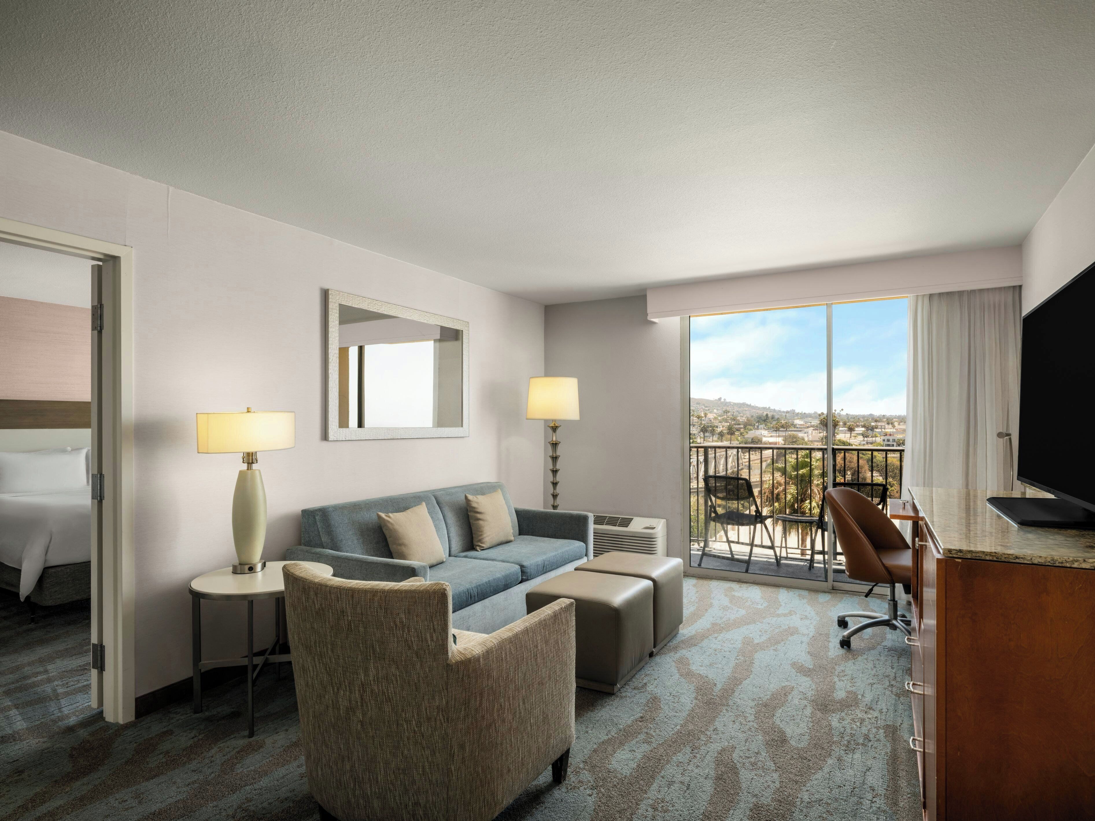 Habitación de hotel con sofá, sillón, escritorio y un amplio ventanal con vistas.
