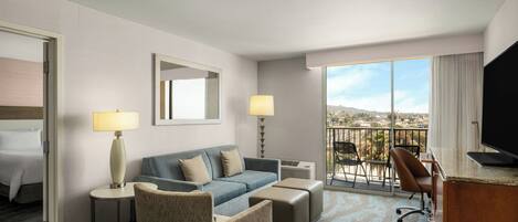 Habitación de hotel con sofá, sillón, escritorio y un amplio ventanal con vistas.