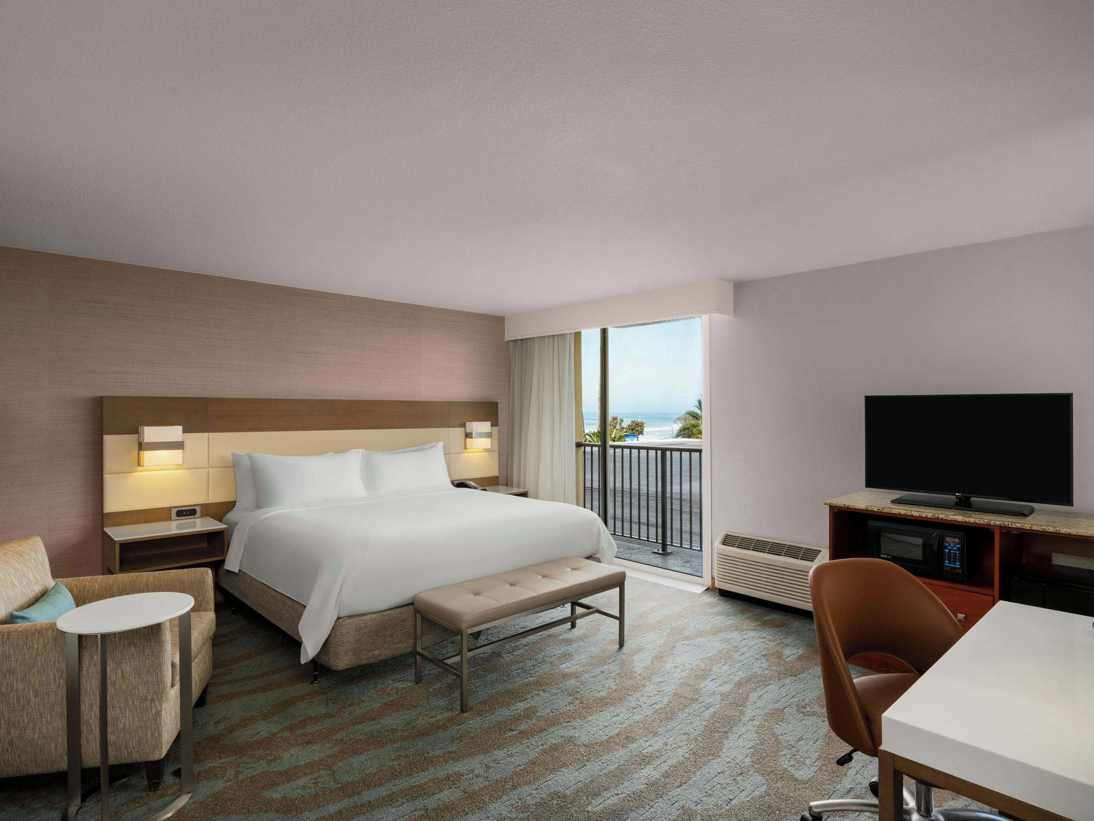 A hotel room with a large bed, a flat-screen TV, a desk, and a view of the ocean.