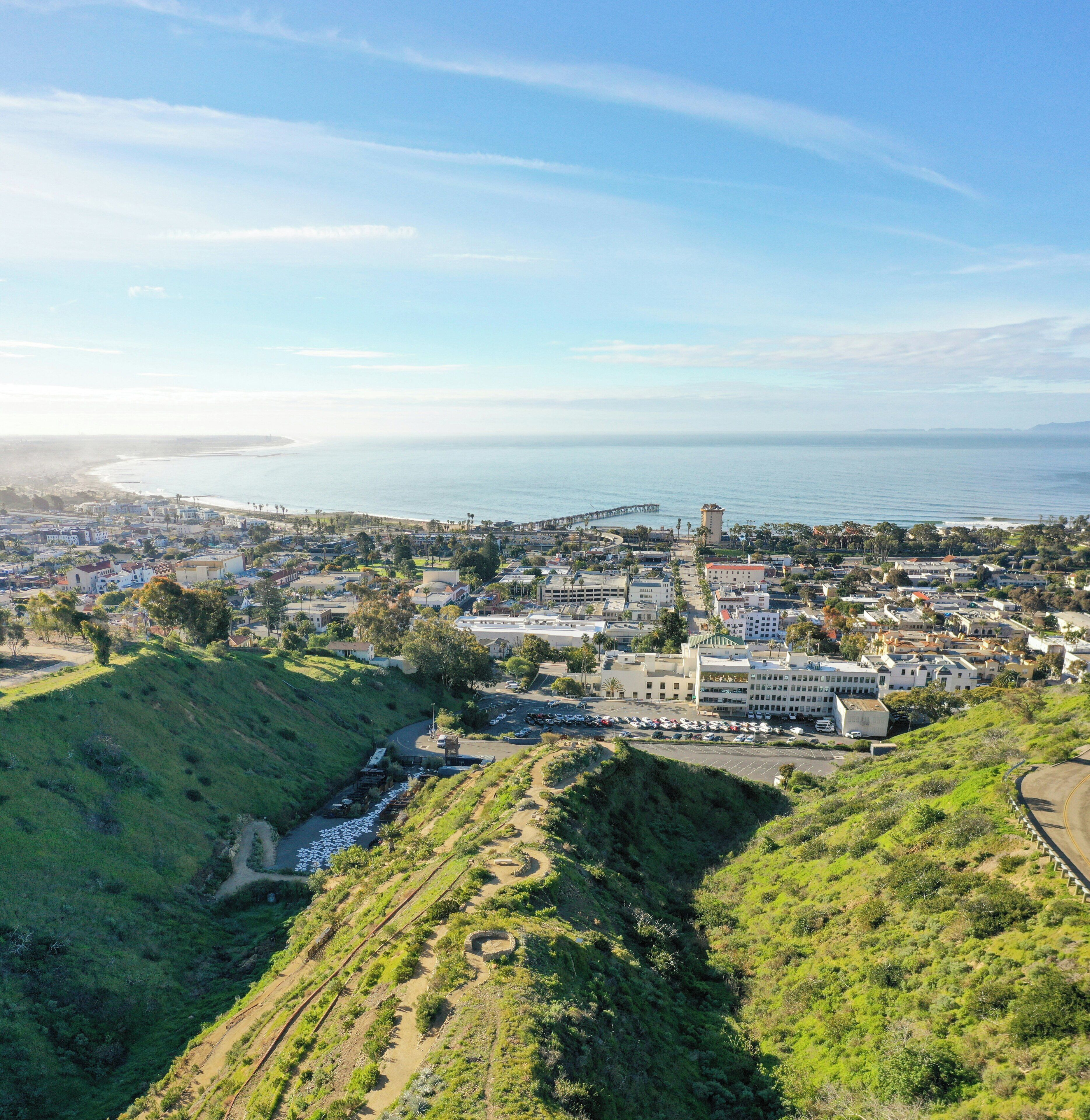 A coastal cityscape with a prominent bridge, a marina, and a hilly terrain with a road curving through it.