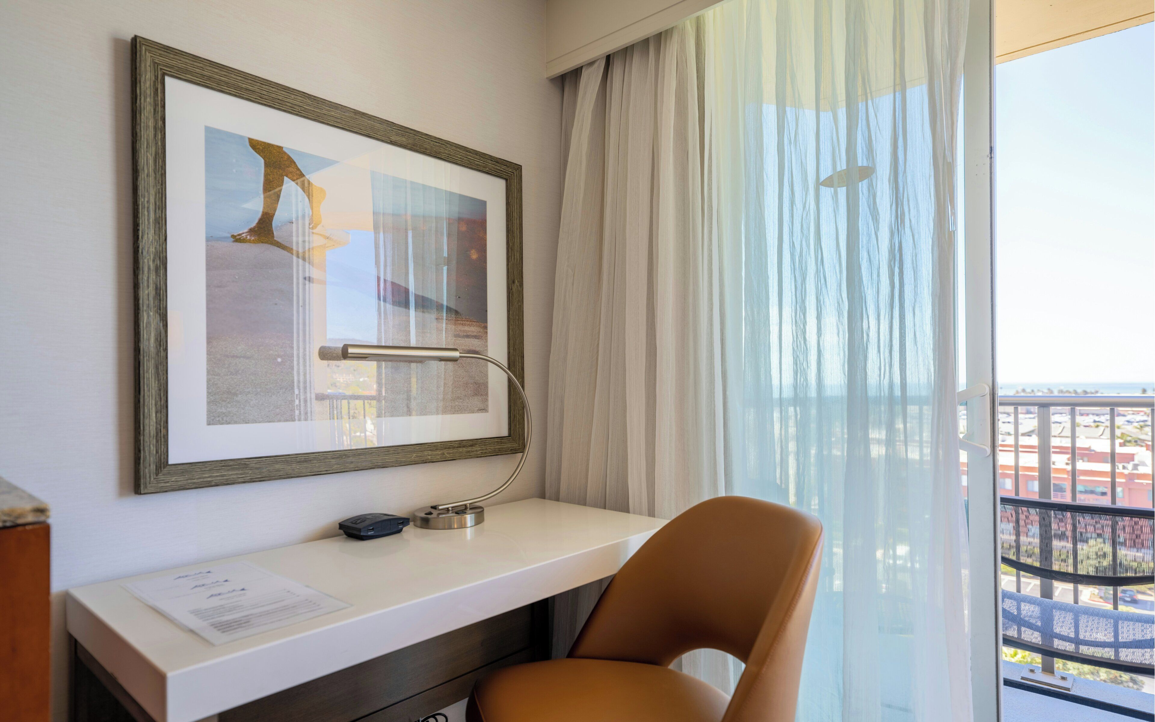 A hotel room with a desk, chair, framed artwork, and a view of a balcony.