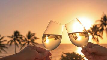 Two hands holding wine glasses with a sunset and palm trees in the background.