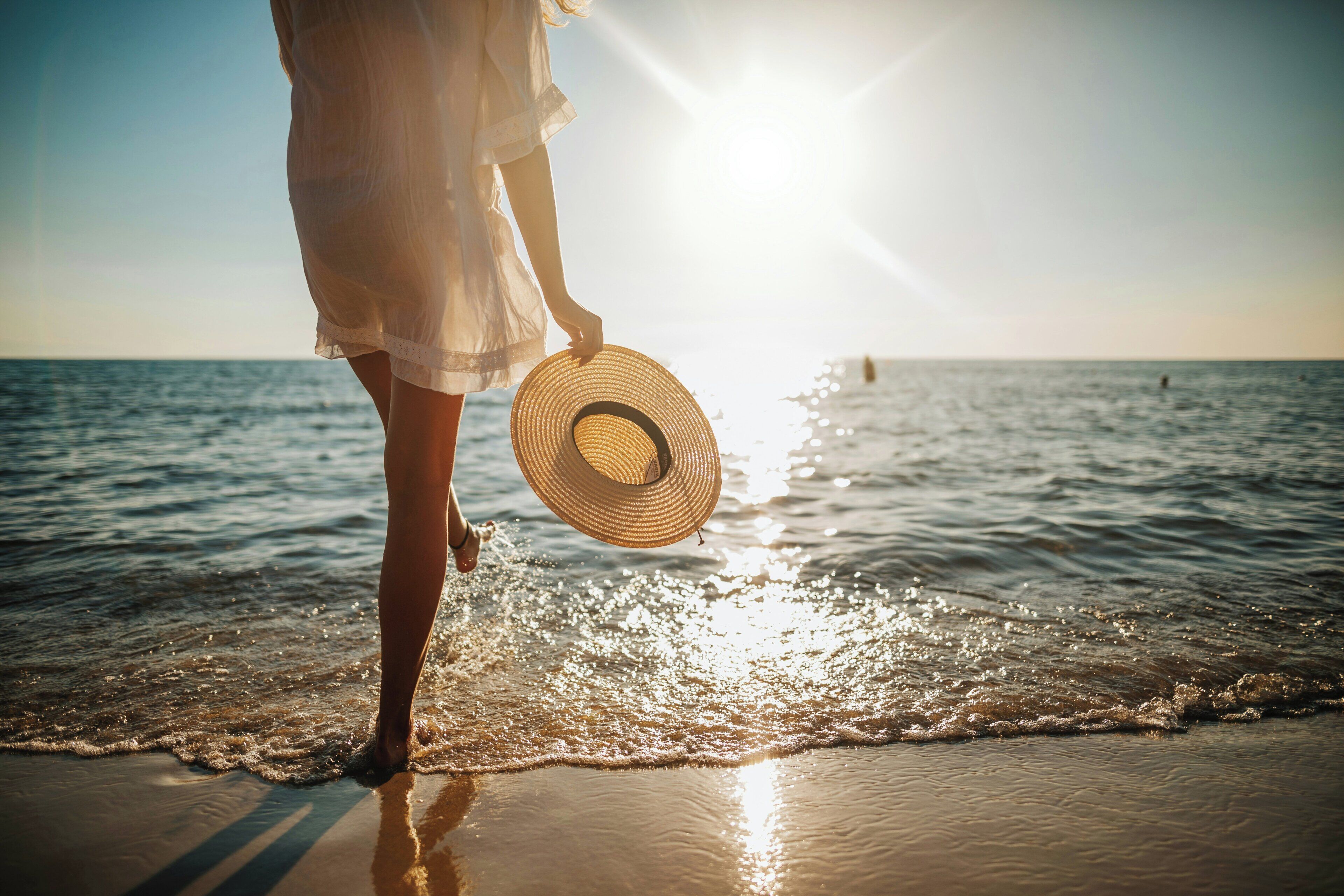 A person standing on a beach holding a hat, with the ocean and a clear sky in the background.