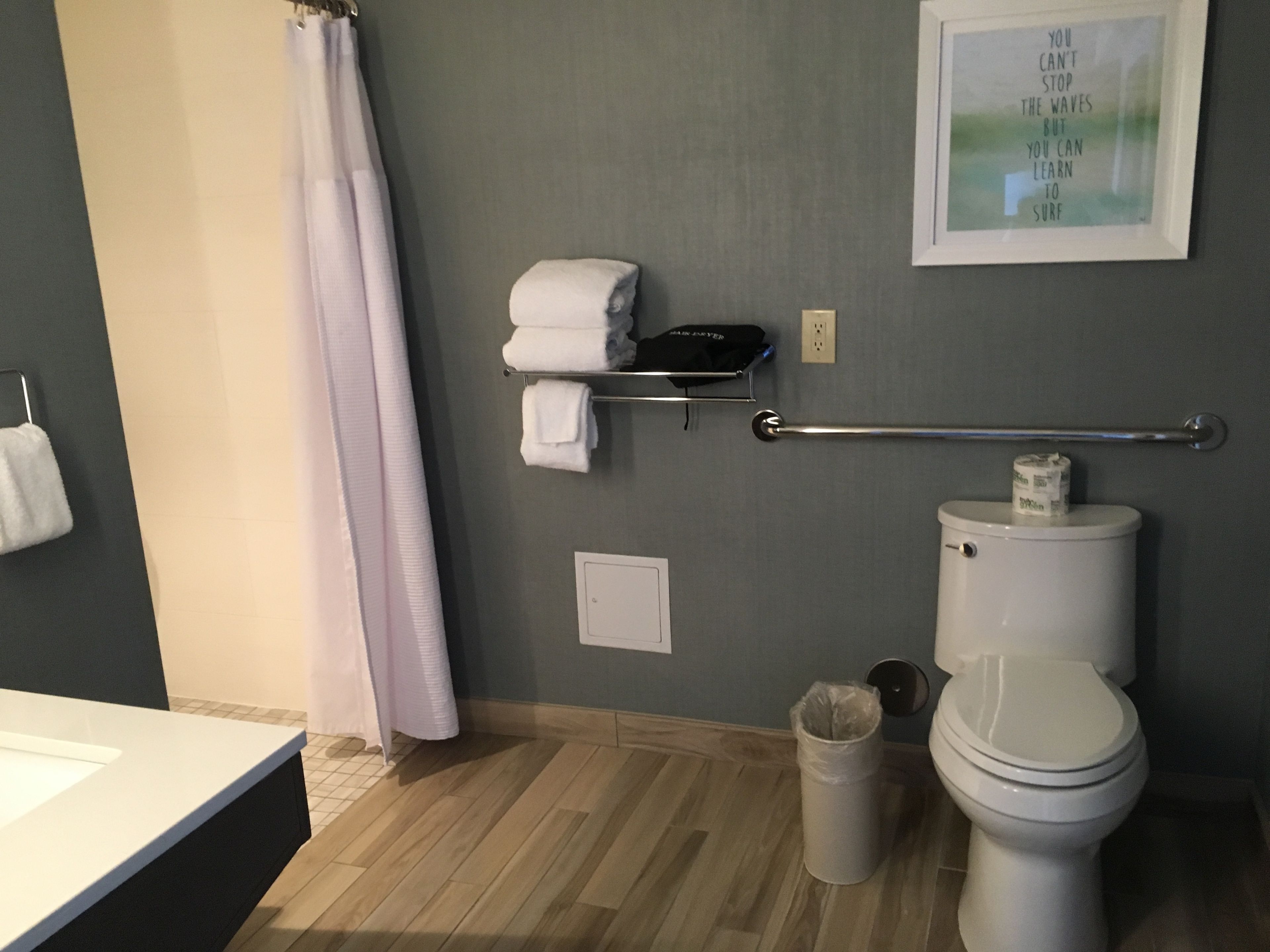 A bathroom with a toilet, a trash can, a towel rack with towels, a shower curtain, and a framed inspirational quote on the wall.