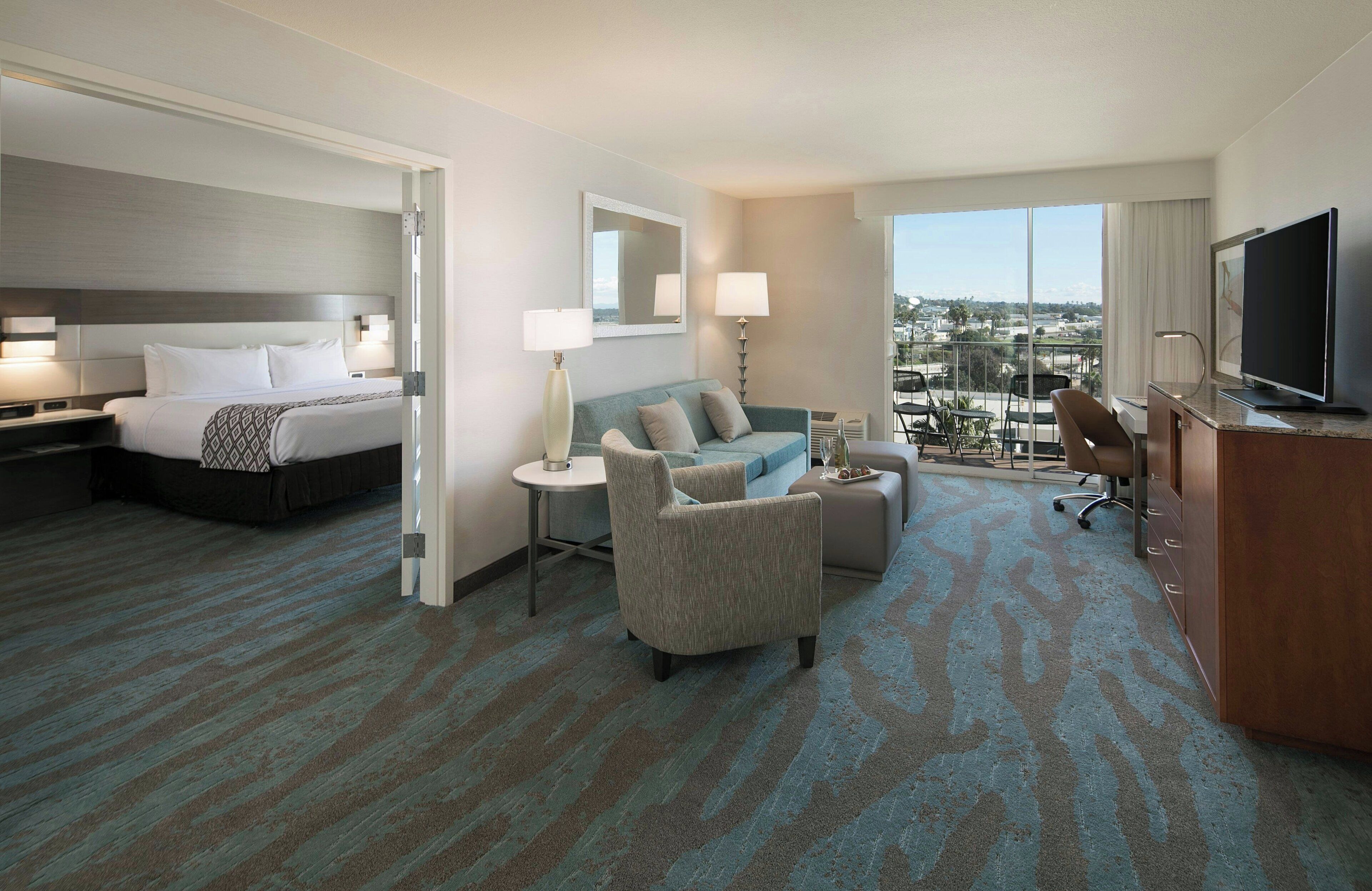 A hotel room with a bed, a sofa, a desk, a TV, and a balcony with a city view.