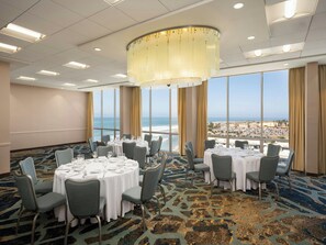 Sala de conferências com mesas redondas, cadeiras e uma grande janela com vista para a praia e a cidade.