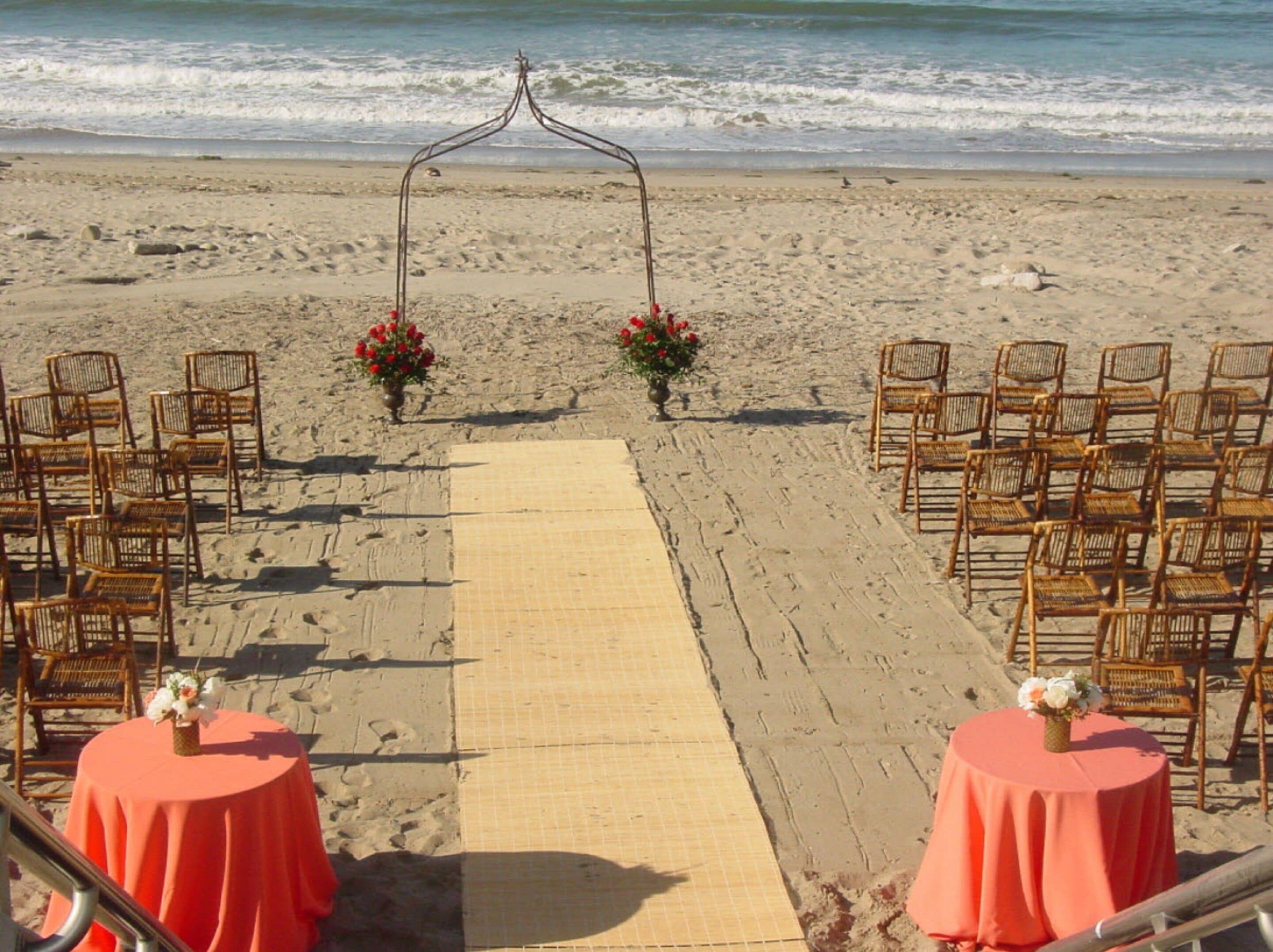 A beach wedding setup with a wooden arch, chairs, and tables with red tablecloths.