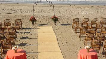 A beach wedding setup with a wooden arch, chairs, and tables with red tablecloths.