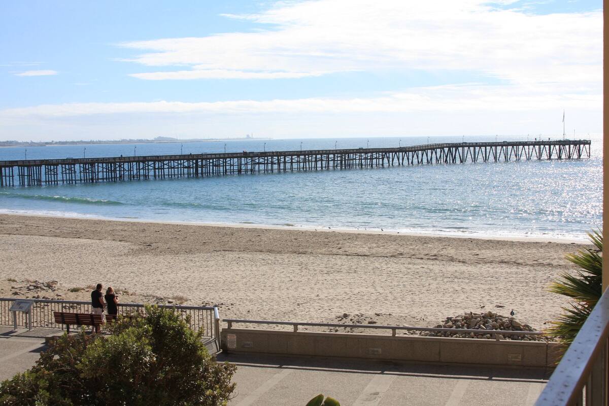 A beach with a pier extending into the water, a sandy shore, and a few people standing by a railing.