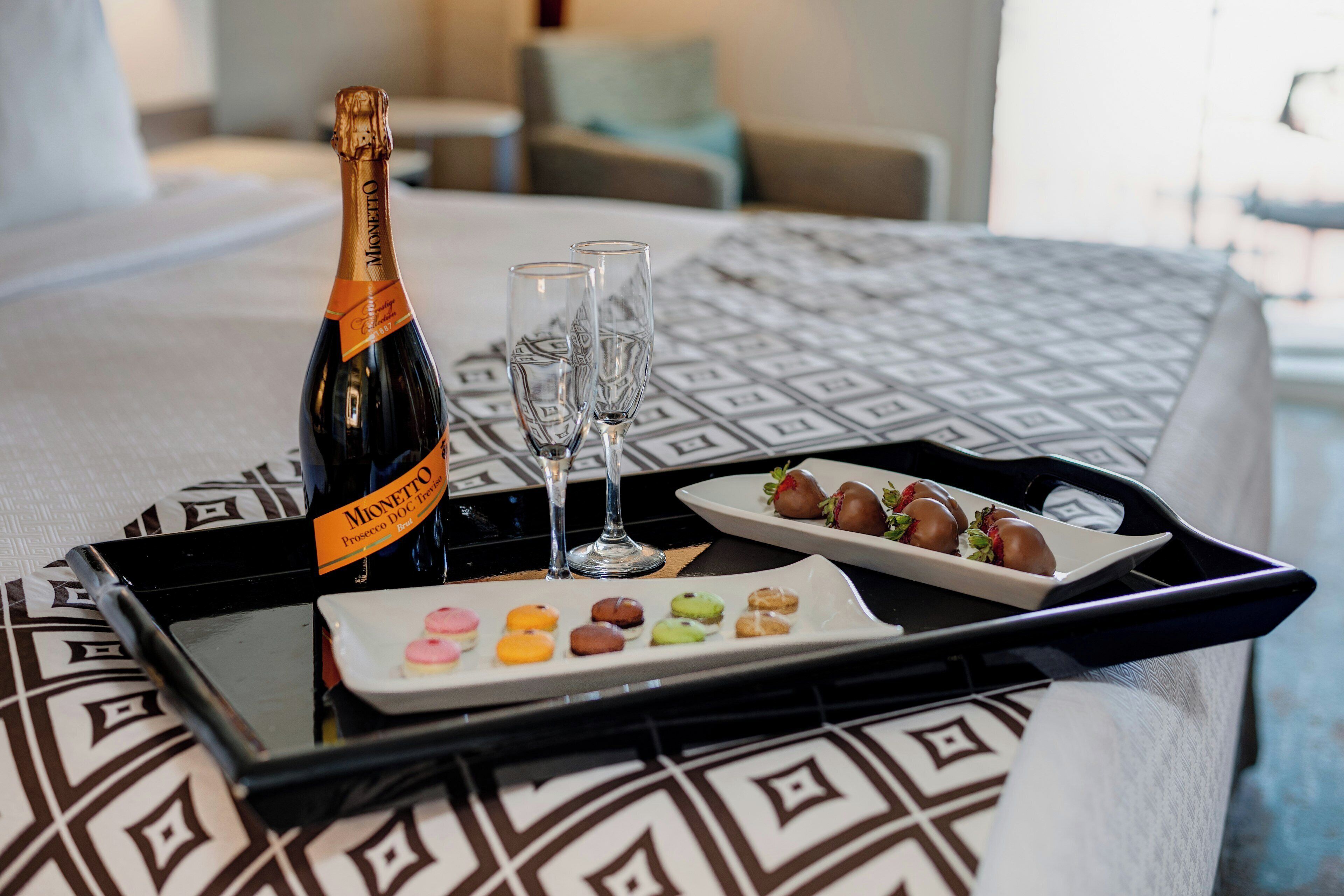 Een fles Mionetto champagne, twee glazen en een dienblad met diverse chocolade en macarons op een bed.