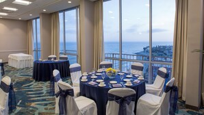 A conference room with a large window overlooking the sea, round tables set for a meeting, and chairs with blue sashes. - Crowne Plaza Ventura Beach by IHG (Ventura)