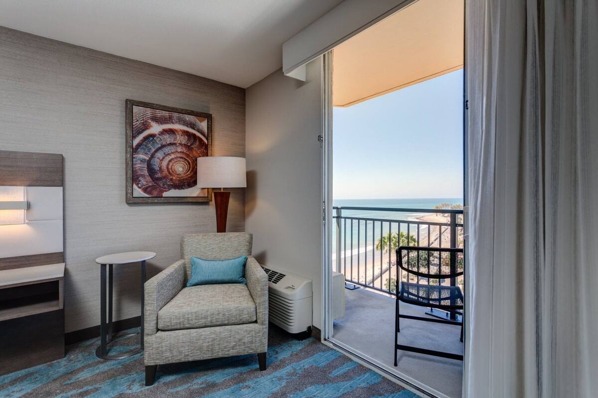 A hotel room with a balcony, a sofa, a small table, a lamp, and a wall art piece.