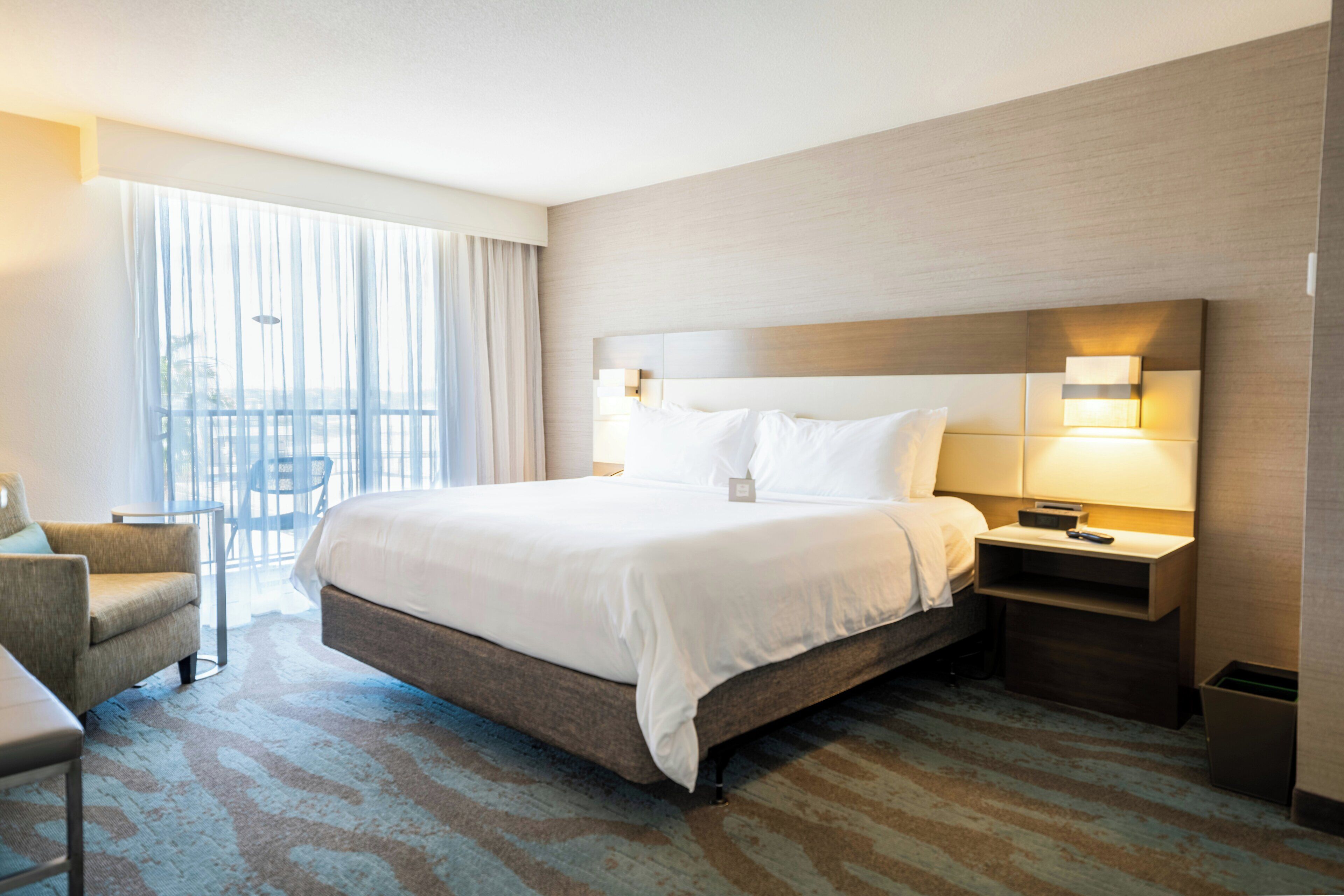 A hotel room with a large bed, a chair, a desk, and a view of the outdoors through a window.