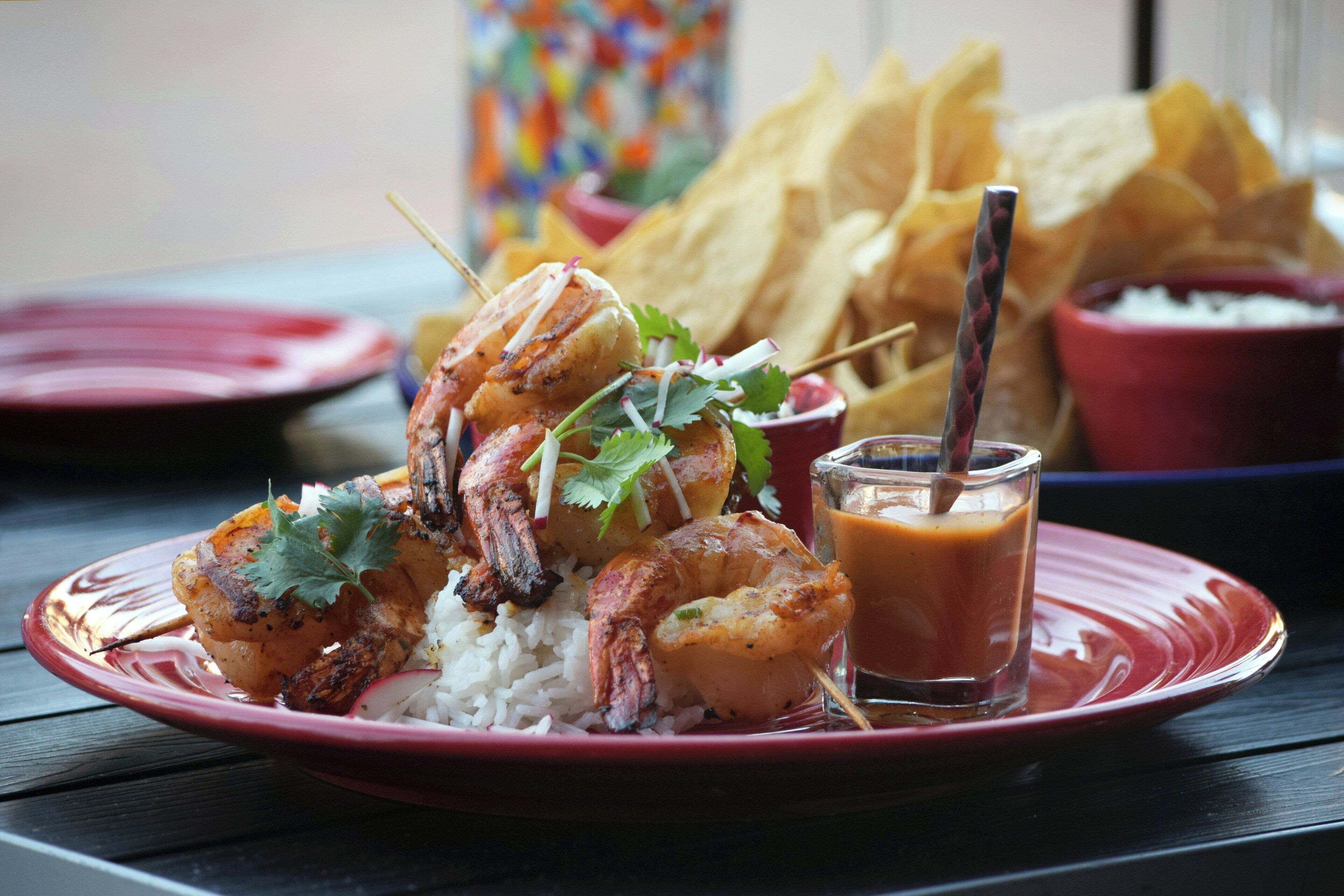 A plate of grilled shrimp and rice, a glass of sauce, and tortilla chips on a table.