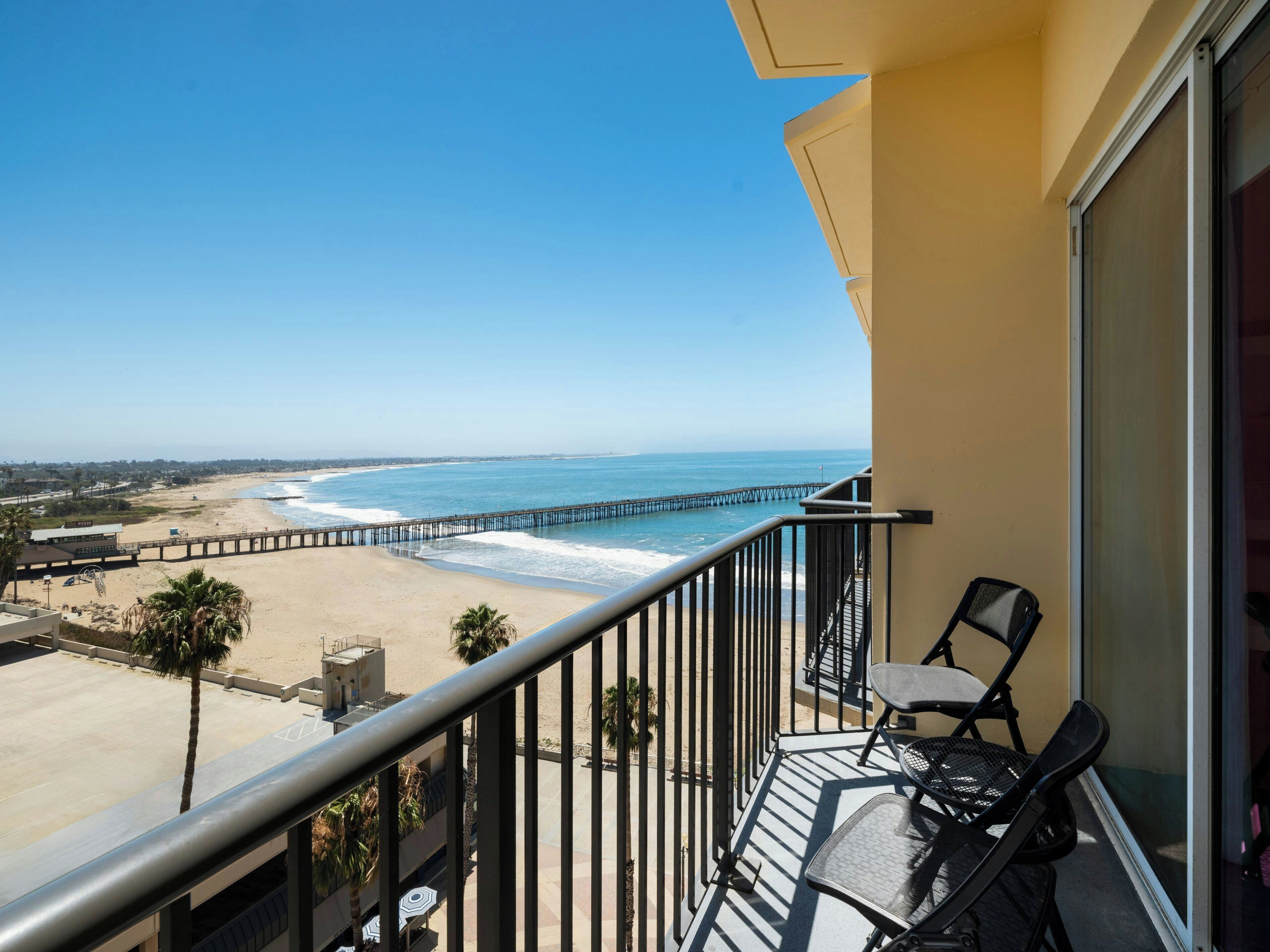 A balcony with a view of a beach and a pier. There are two chairs and a table on the balcony.