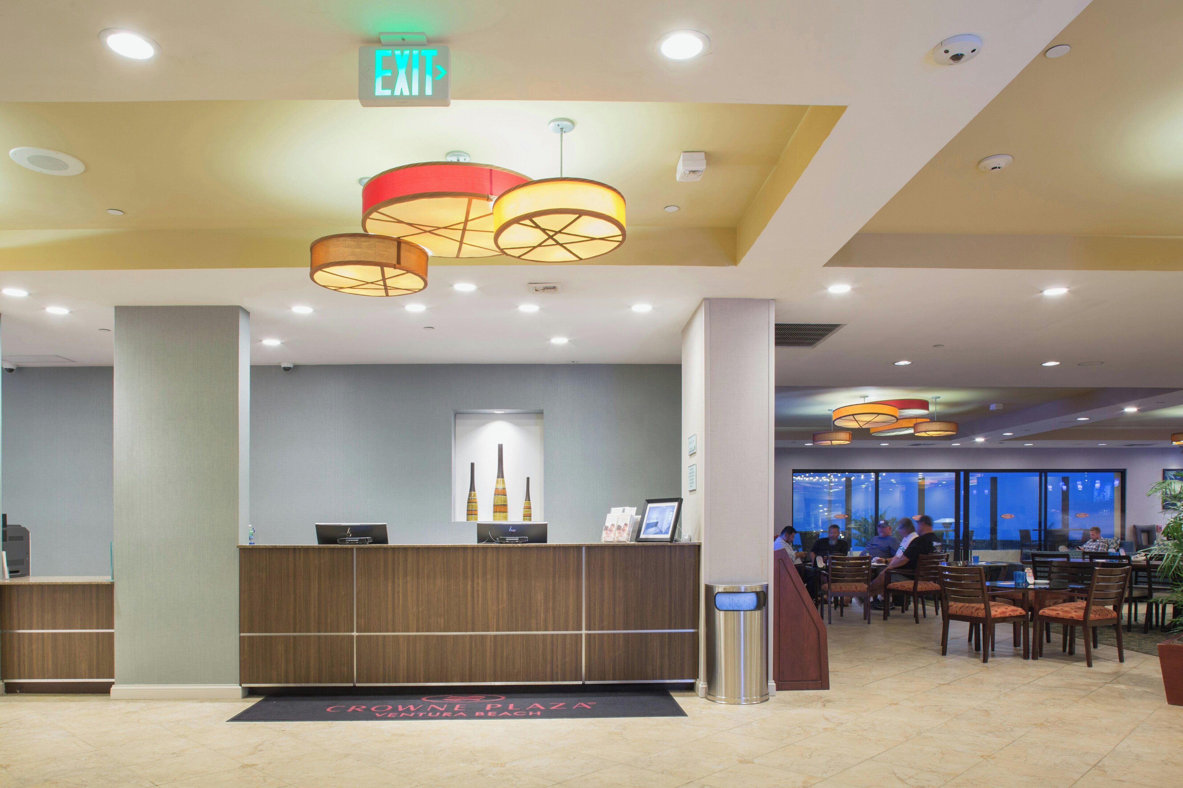 A hotel lobby with a reception desk, a digital screen, and a view of the dining area.