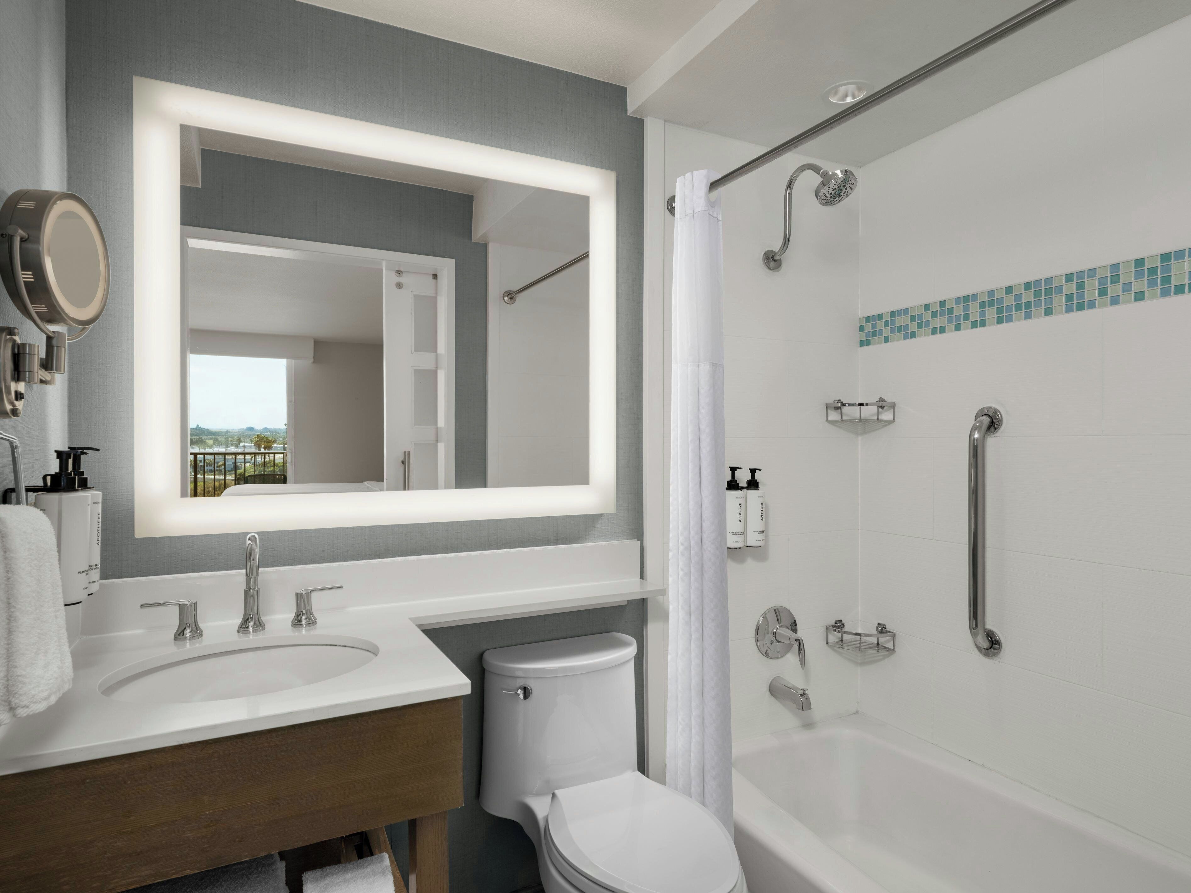A modern bathroom with a large mirror, dual sinks, a toilet, and a walk-in shower with grab bars.