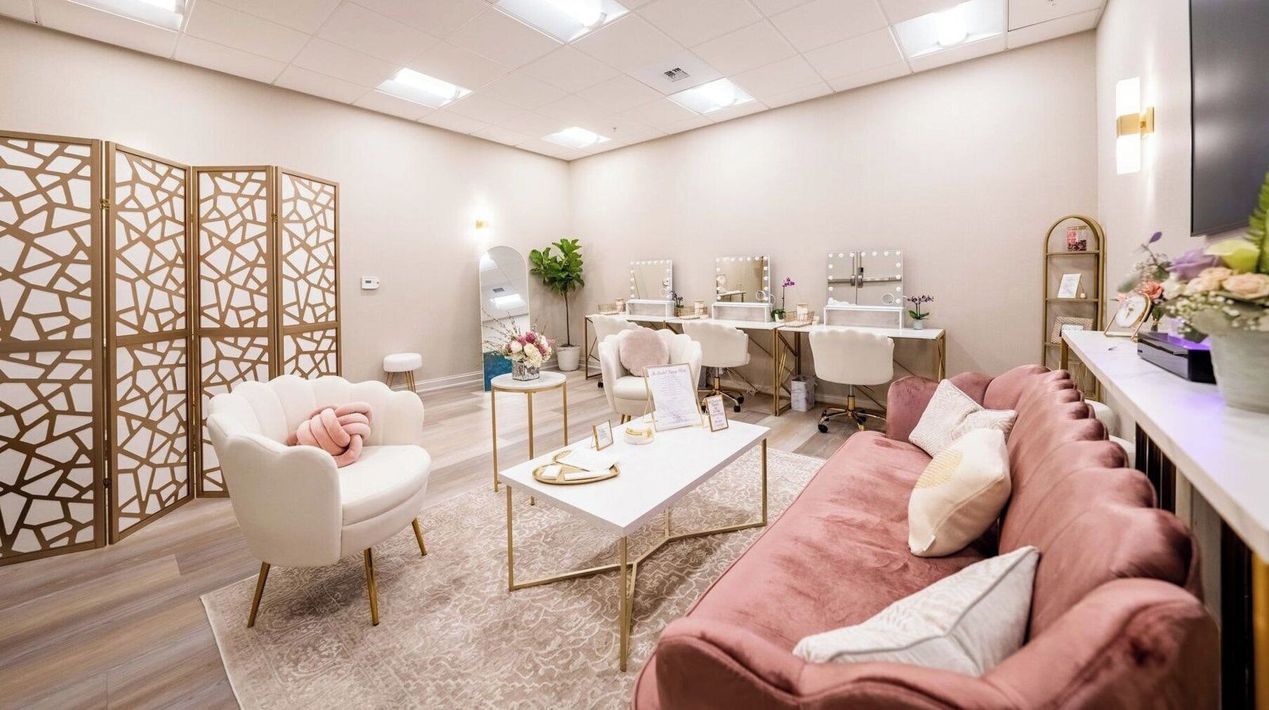 Un salon bien éclairé, avec un canapé blanc, un divan rose et un coin coiffeuse agrémenté de miroirs et de luminaires.