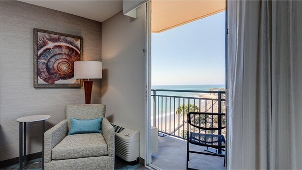 A hotel room with a view of the beach, a chair, a table, a lamp, and a large artwork on the wall. - Crowne Plaza Ventura Beach by IHG (Ventura)