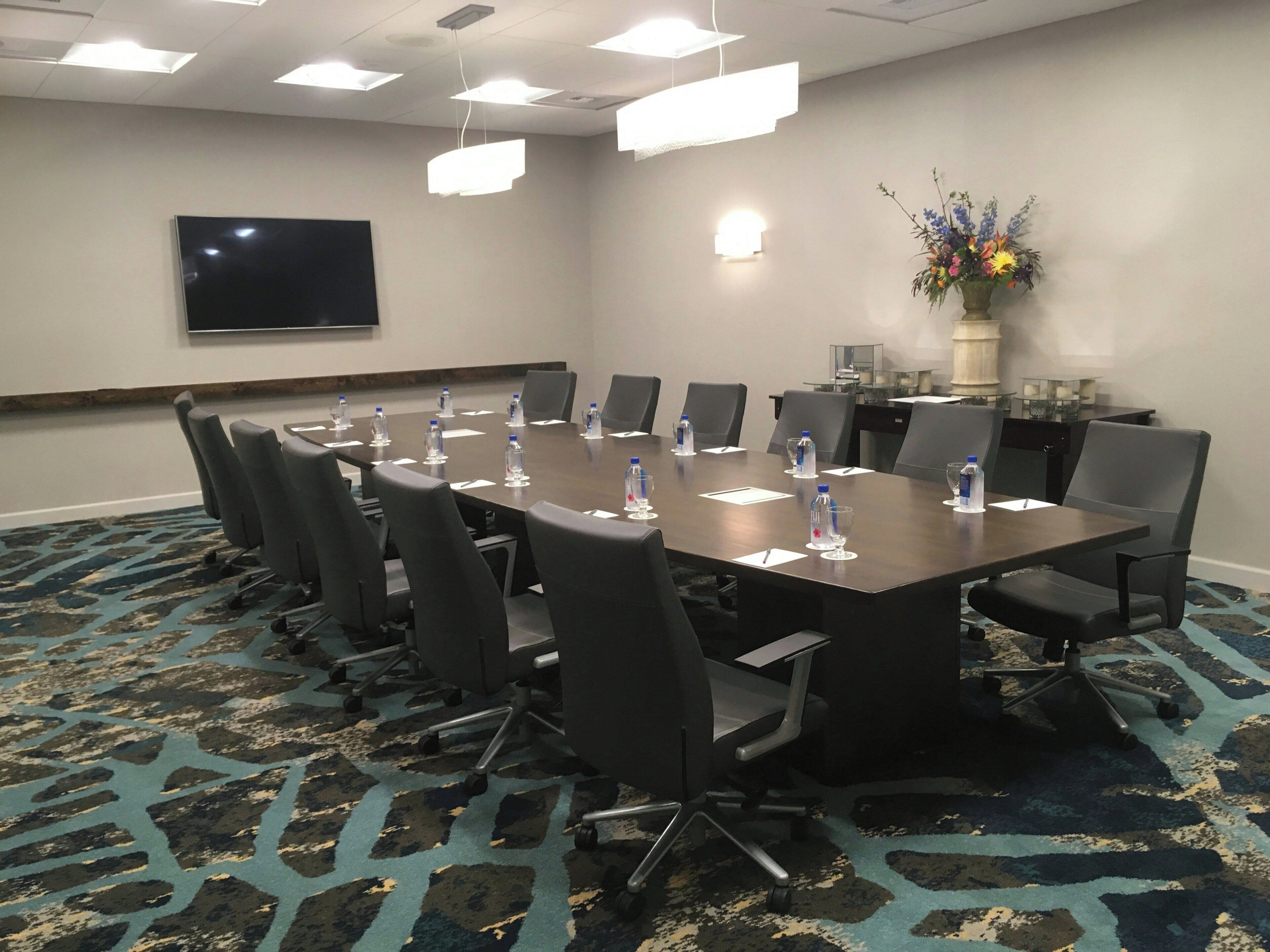 Une salle de conférence avec une longue table, des chaises, des bouteilles d’eau et un téléviseur à écran plat fixé au mur.