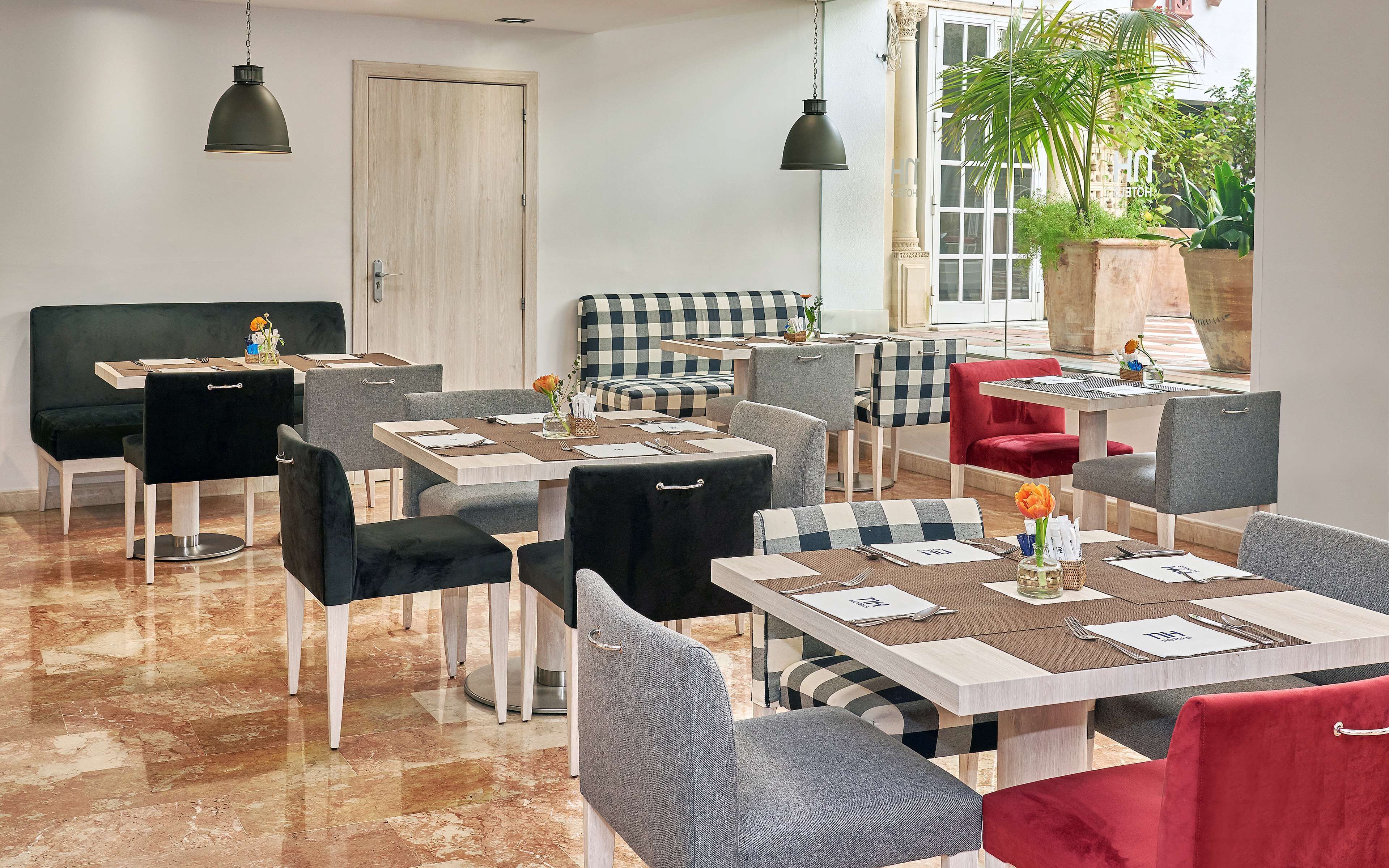 A modern dining area with multiple tables and chairs, a checkered sofa, and a large window with a view of greenery.