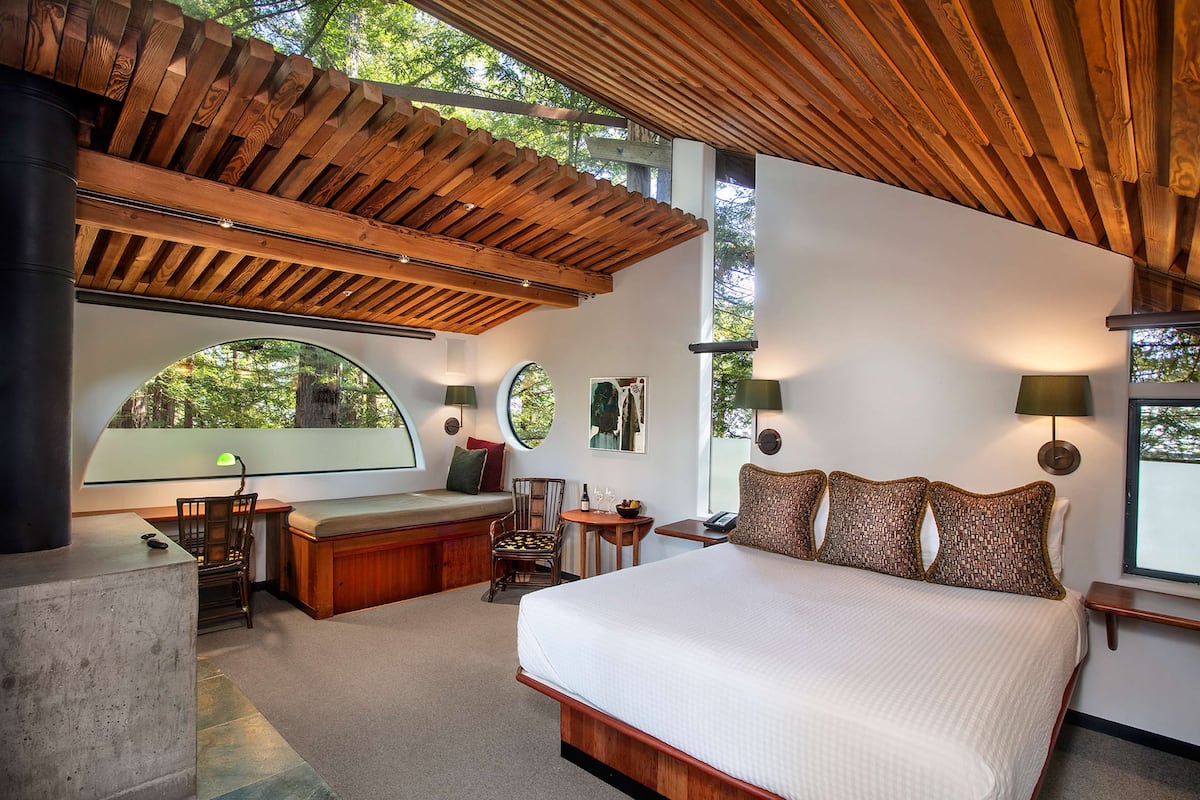 A bedroom with a large bed, wooden ceiling, and a view of trees through a window.