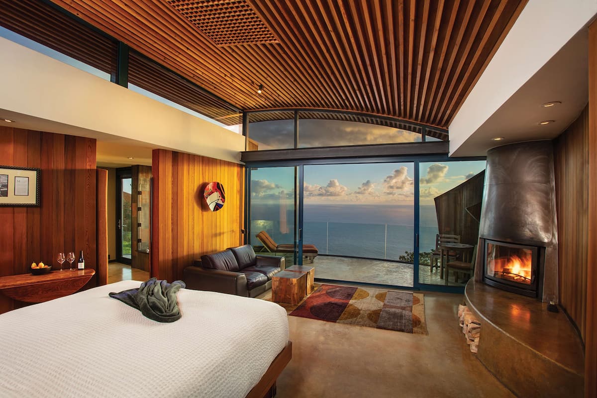 A modern bedroom with a large bed, a fireplace, and a balcony with a view of the ocean.