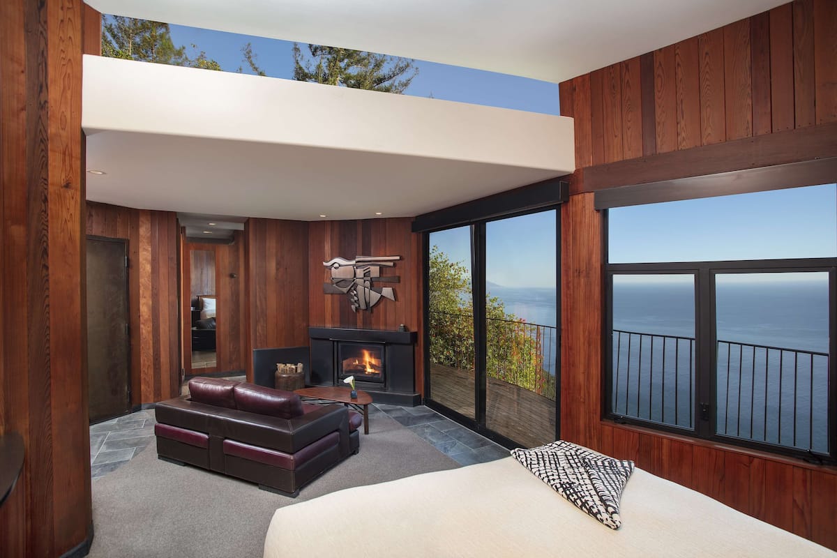 A modern bedroom with a fireplace, a large window with a view of the ocean, and wooden paneling.