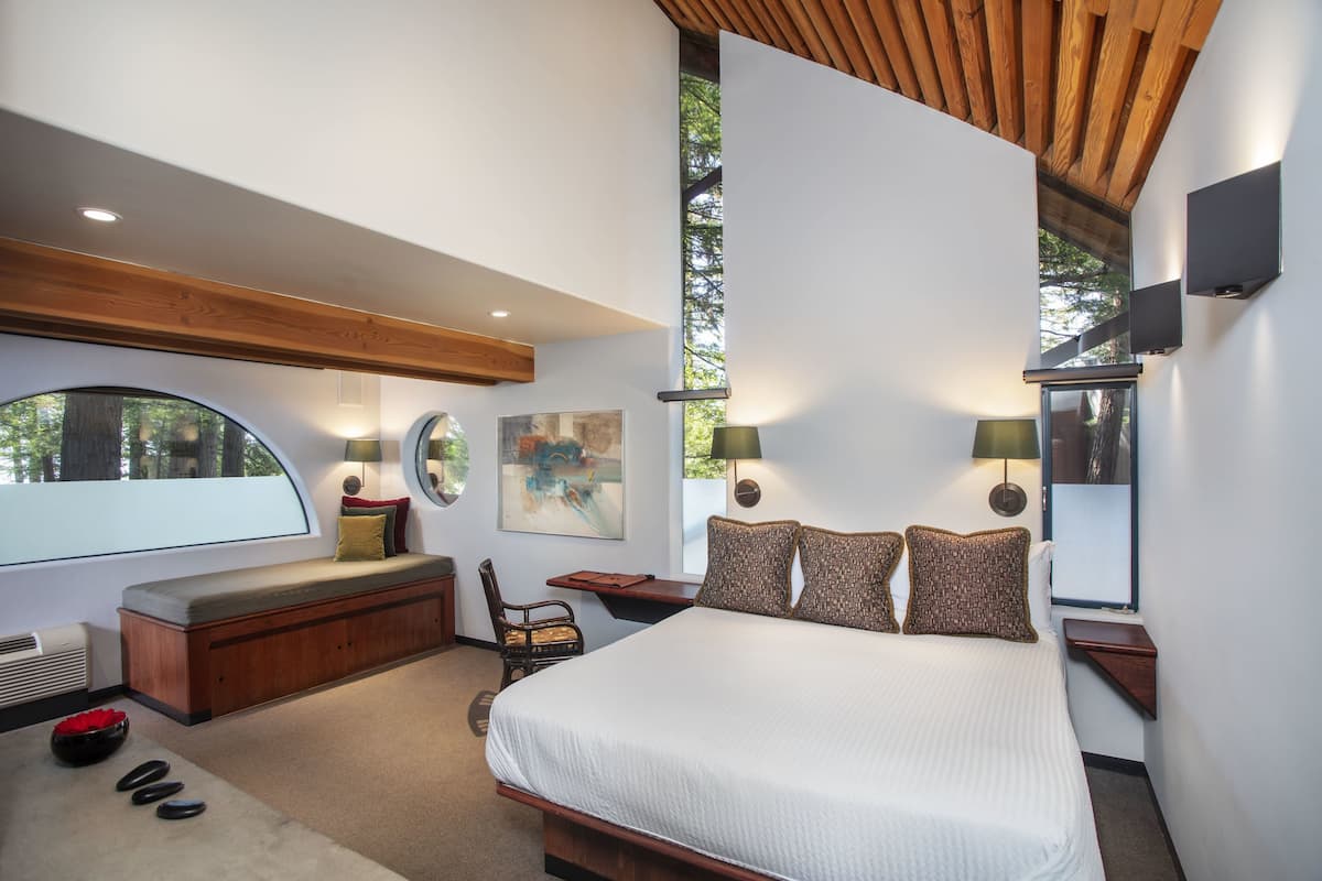 A modern bedroom with a large bed, a bench, a desk, and a large window.
