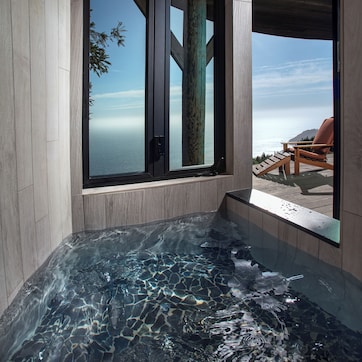 A modern spa area with a glass-enclosed hot tub, wooden deck, and a view of the ocean.