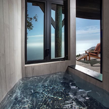 A modern spa area with a glass-enclosed hot tub, wooden deck, and a view of the ocean.