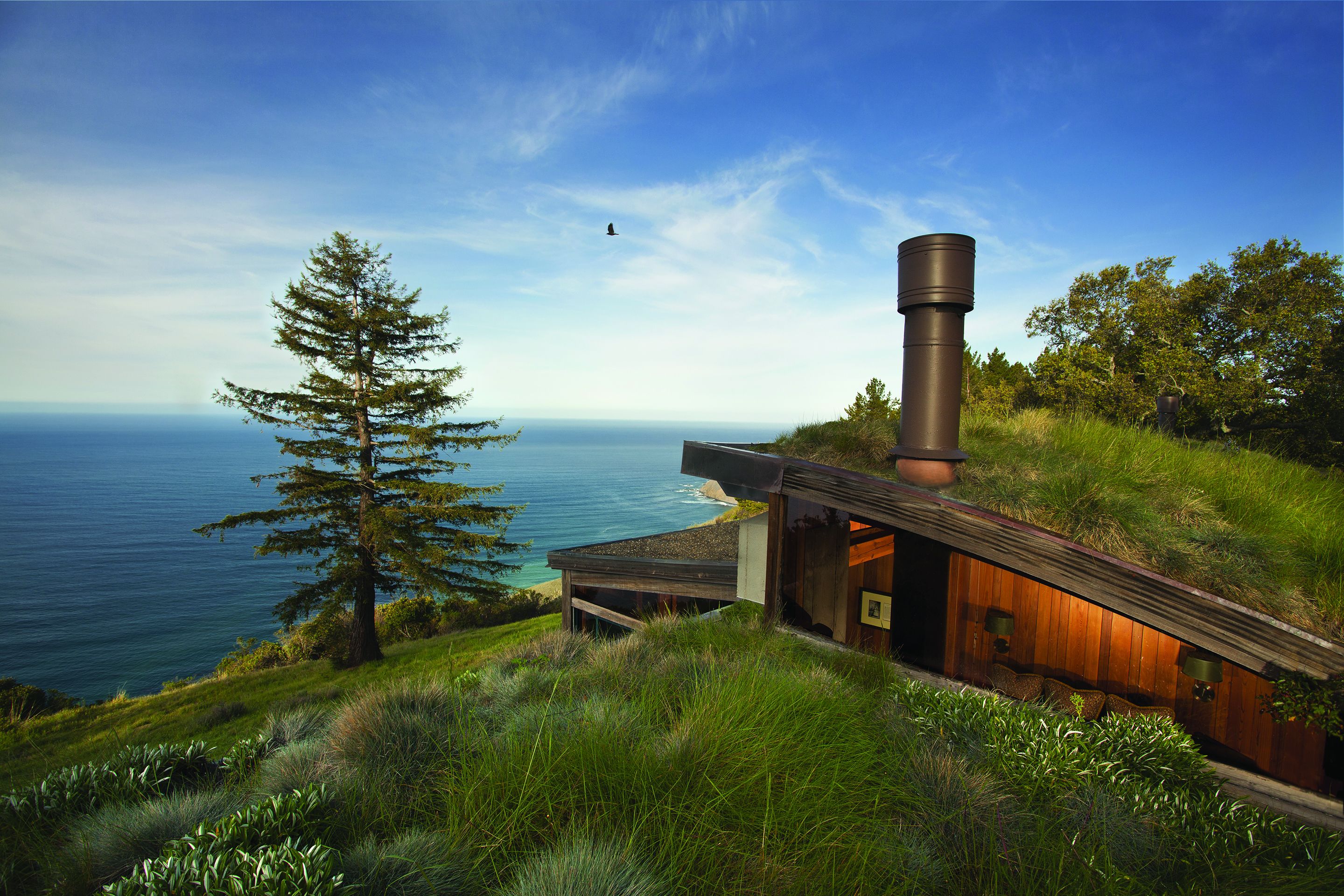 A modern house with a large chimney, situated on a grassy hill overlooking a body of water.