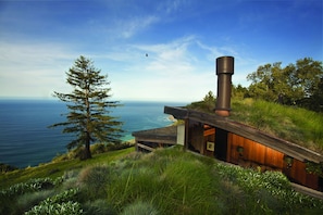 A modern house with a large chimney, situated on a grassy hill overlooking a body of water.