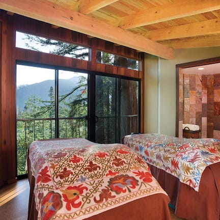 A bedroom with two beds, large windows, a view of mountains, and a stone wall.