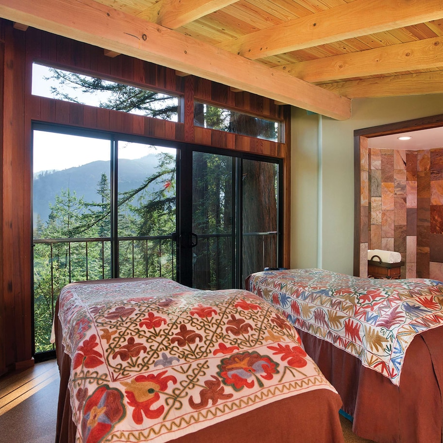 A bedroom with two beds, large windows, a view of mountains, and a stone wall.