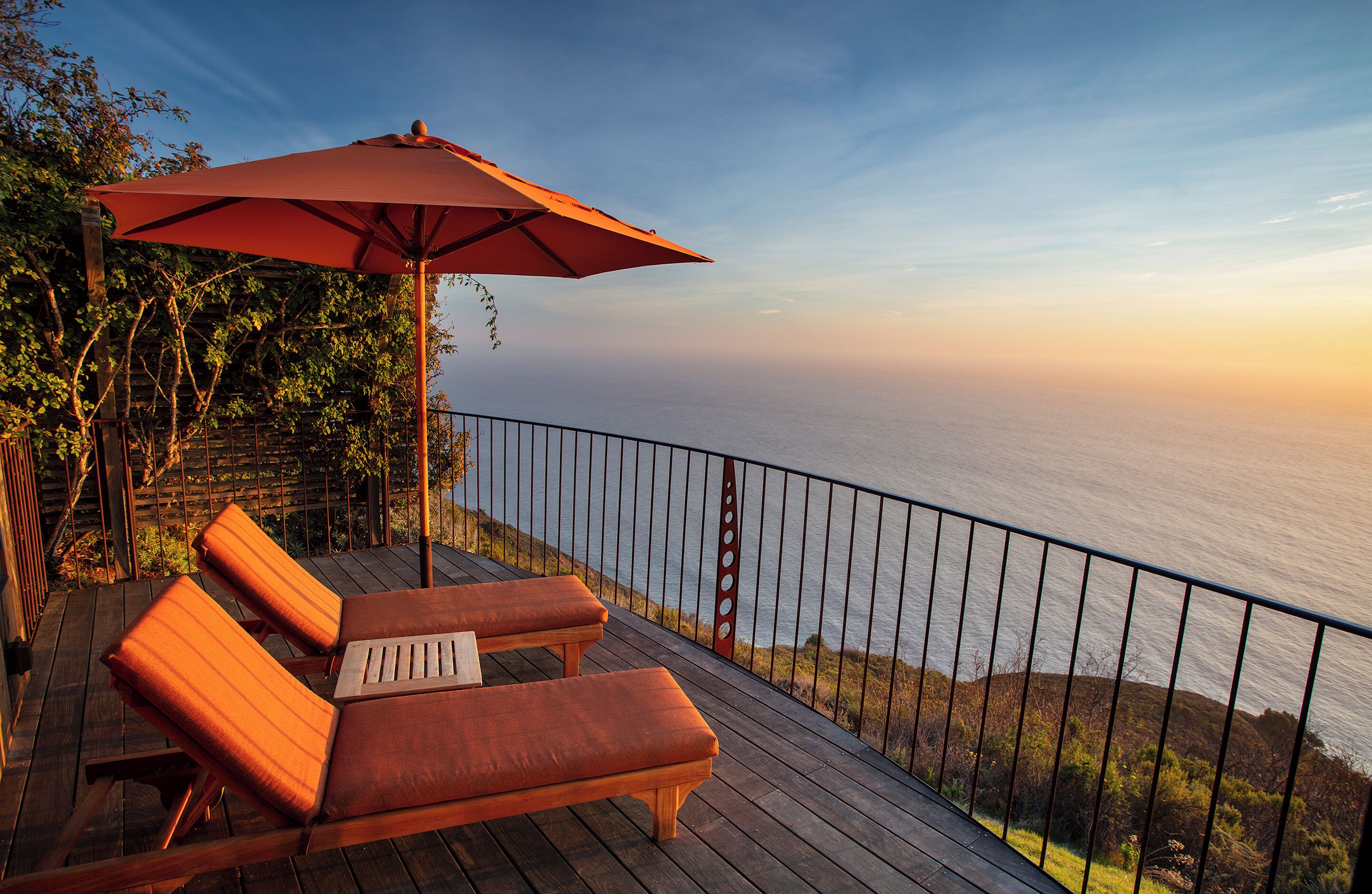 Um deck de madeira com duas cadeiras de descanso e um guarda-sol vermelho, com vista para um corpo de água.