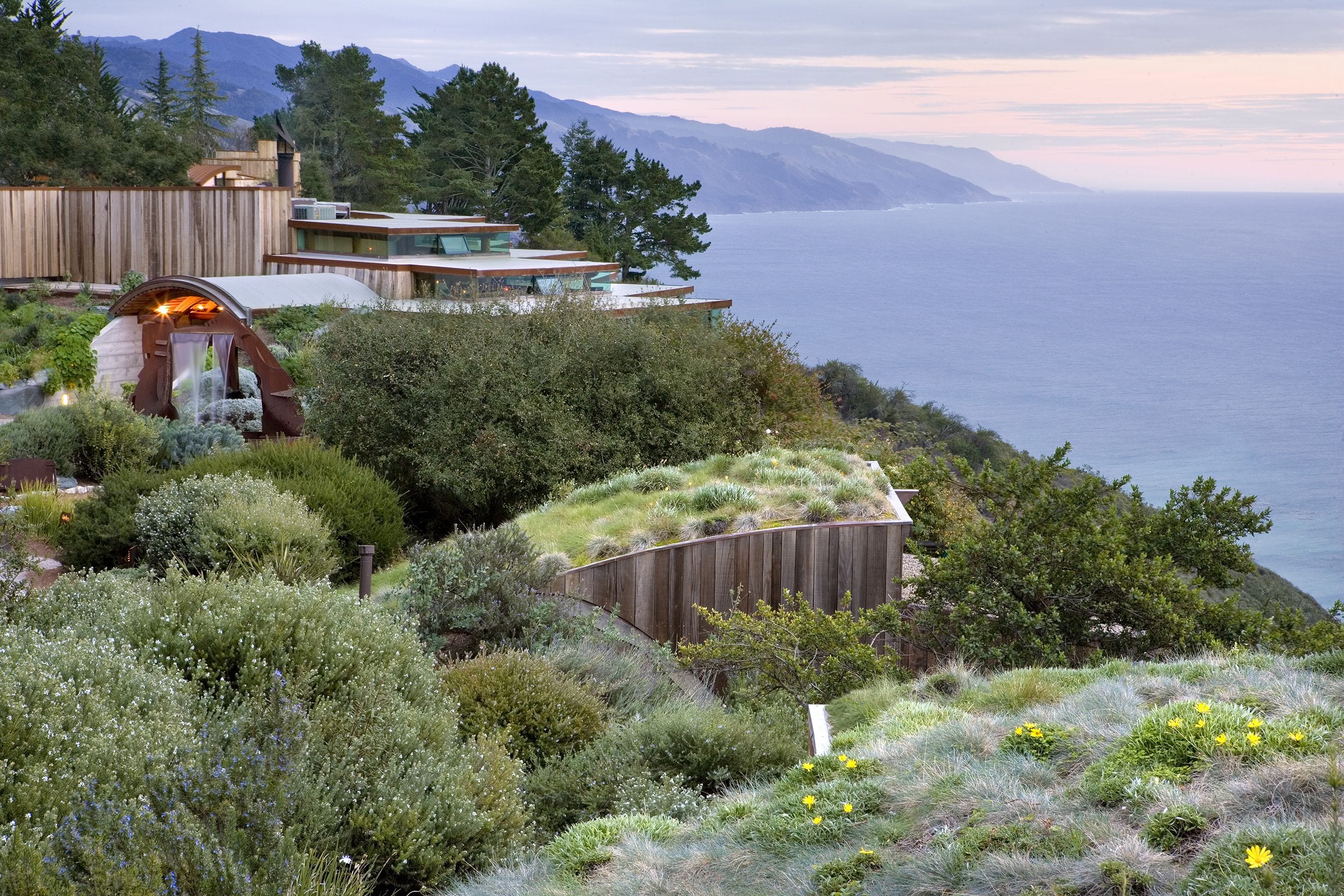 A modern house with a unique green roof, surrounded by lush vegetation and overlooking a body of water.