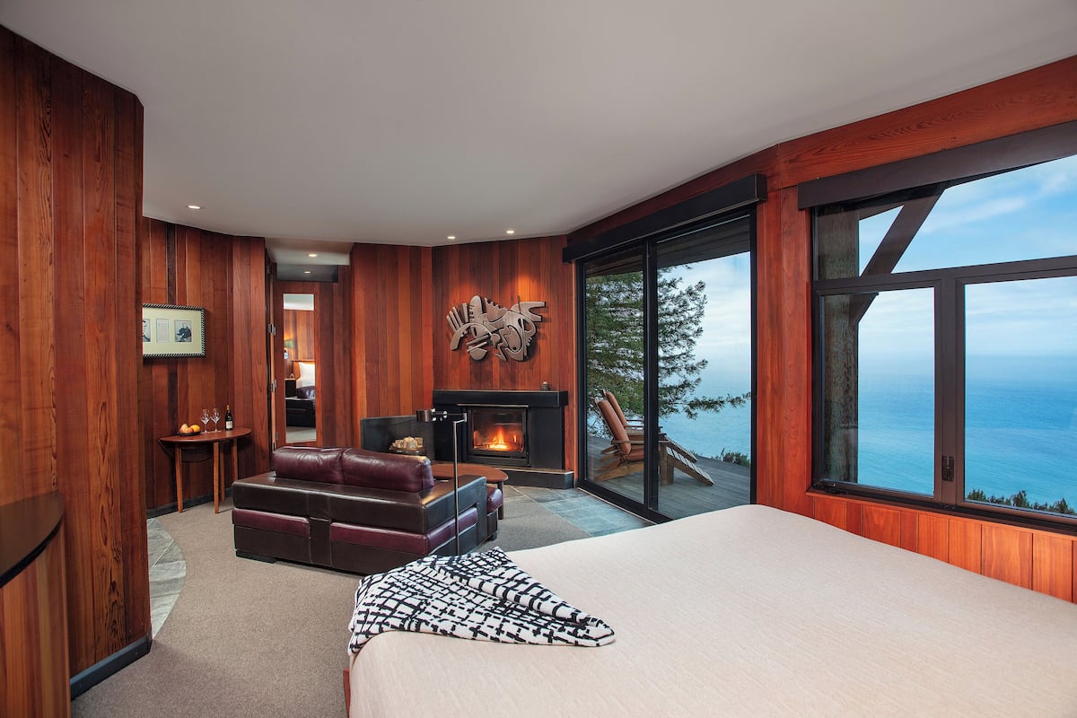 A modern bedroom with a large bed, a fireplace, a leather sofa, and a view of the ocean.