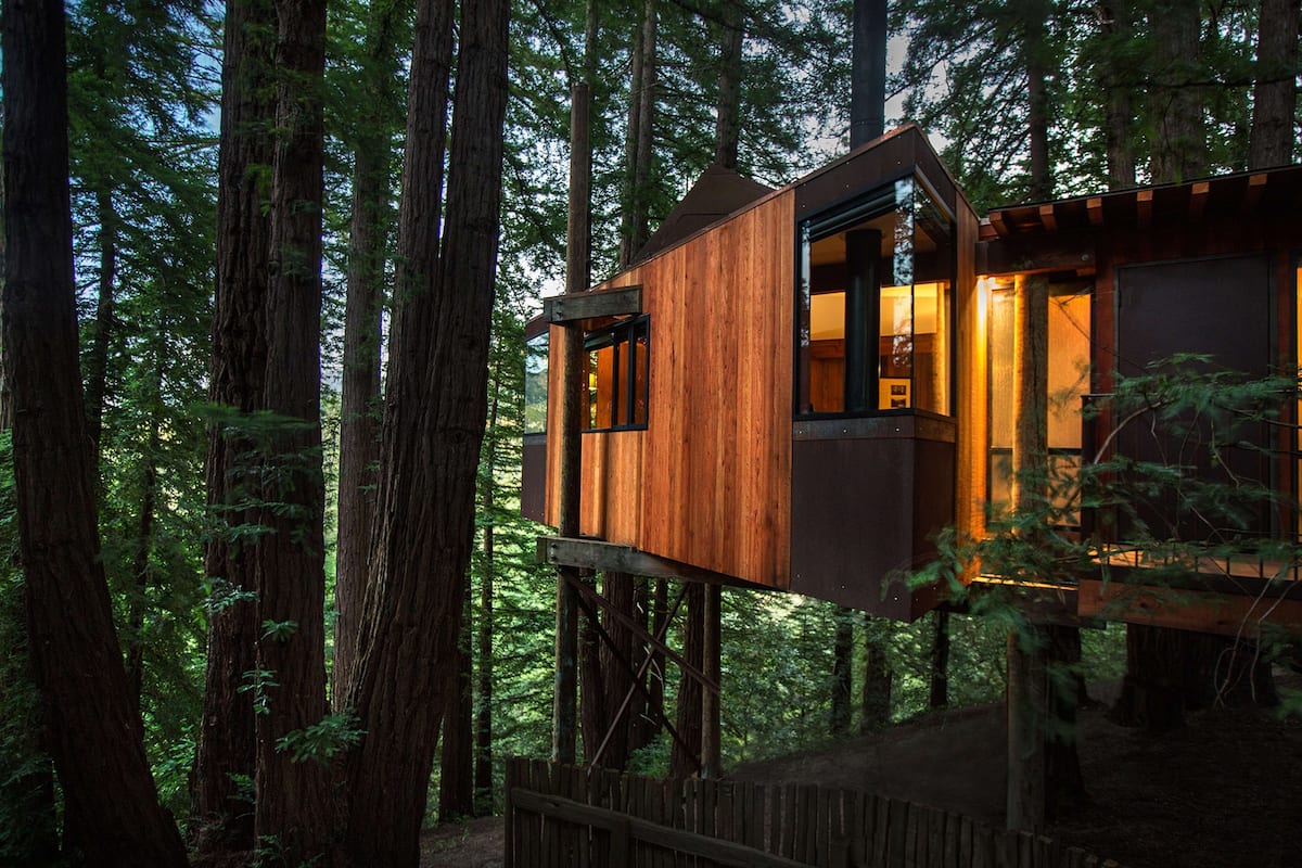 A wooden treehouse with a deck, surrounded by tall trees.