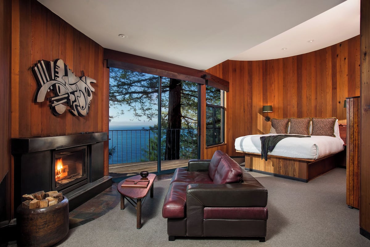 A modern bedroom with a fireplace, a large bed, a sofa, and a view of the outdoors.