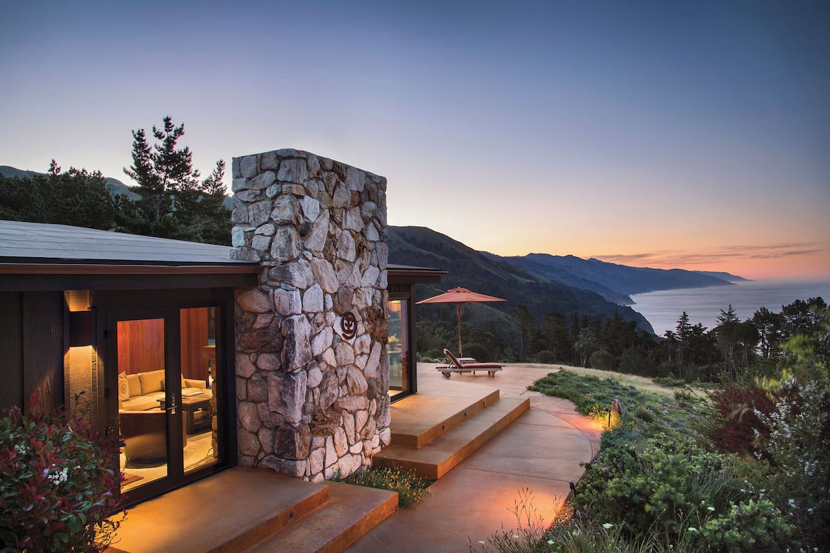 A stone-clad building with a patio and outdoor seating area, overlooking a mountainous landscape.
