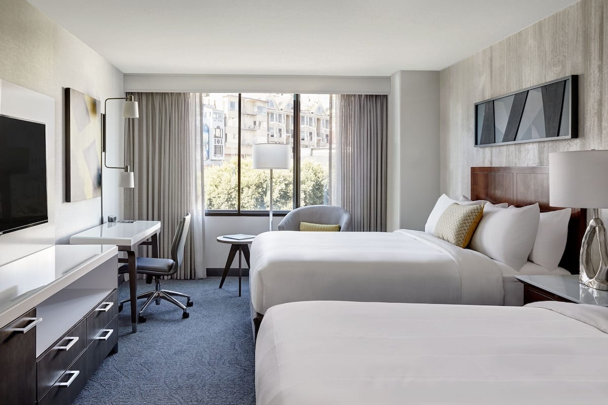 A hotel room with two beds, a desk, a chair, a TV, and a window with curtains.