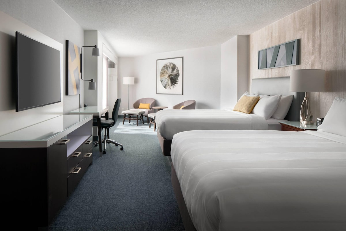 A hotel room with two beds, a desk, a chair, a TV, and artwork on the walls.