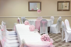A banquet hall with round tables, white chairs with pink sashes, and floral centerpieces. - Holiday Inn Leamington Spa - Warwick by IHG (Warwick)